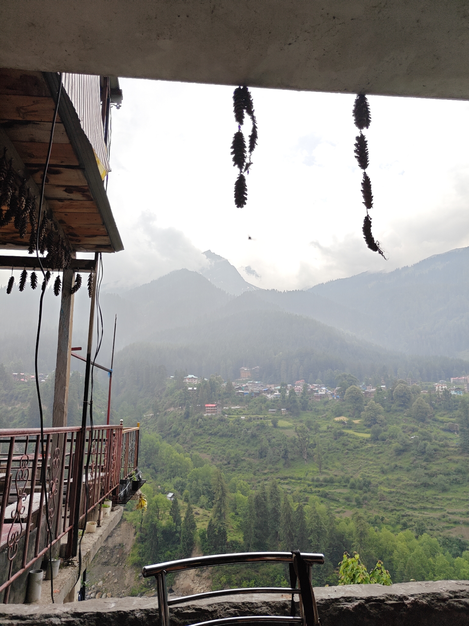 The image captures a panoramic view of a mountainous valley, seen from a high vantage point, likely a balcony or terrace in Pulga, India.
In the immediate foreground, architectural elements frame the scene. A concrete slab forms the top and bottom edges of the frame. On the left, wooden eaves extend from a building, adorned with multiple strings of dark, elongated, pinecone-like decorations. Similar decorative strings hang from the upper right concrete overhang. A reddish-brown metal railing with intricate scrollwork runs along the left, part of a balcony that overlooks a steep drop. Several potted plants are placed along this railing. A silver metallic chair back or railing is partially visible in the bottom right corner. Wires or cables run vertically along the left side.
The midground reveals a lush, verdant valley covered in dense green forests, with numerous trees, including conifers. A small town or village is scattered across the valley slopes, comprising many buildings with roofs of various colors like red, blue, and grey, nestled amidst the greenery. Some terraced or cultivated land is also visible on the slopes.
In the background, majestic mountains rise, partially obscured by heavy mist and clouds, creating a hazy atmosphere. The sky is overcast, indicating a cloudy daytime, possibly cool and humid weather. There is no visible human activity, nor any discernible text. The scene evokes a sense of peace and natural grandeur, characteristic of the Himalayan region.
