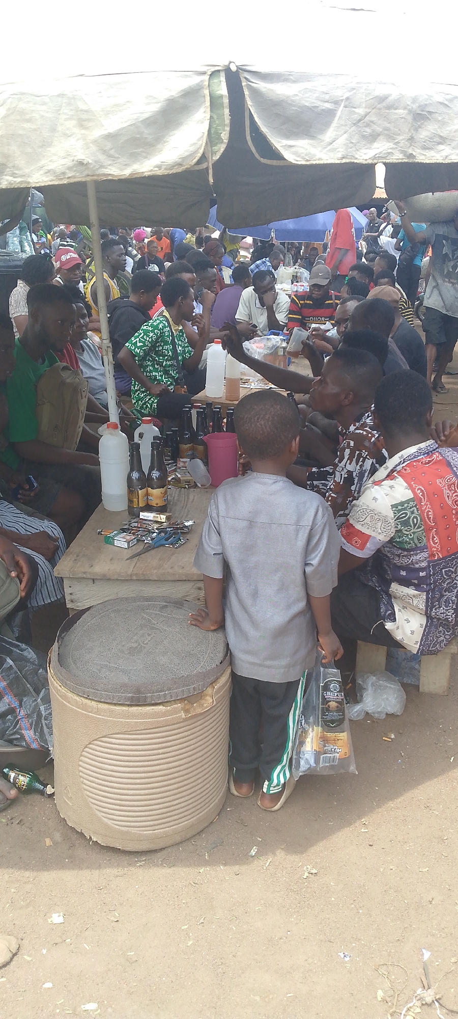 The image depicts a busy outdoor gathering, possibly a market or a social event, under a large, makeshift canopy. The scene is crowded with people, predominantly men, dressed in casual attire. A low wooden table is central to the activity, laden with bottles of beverages, packets of cigarettes, and what appear to be some consumer goods in plastic packaging. Several individuals are seated around the table, engaged in conversation and consumption. In the foreground, a young boy in a grey shirt and dark pants stands facing away from the camera, holding a plastic bag with items. The ground is dusty, suggesting a dry environment. The overall atmosphere appears informal and communal. The provided hints suggest this scene is located in Tor-Donga, Nigeria, a place known for its vibrant markets and community gatherings.