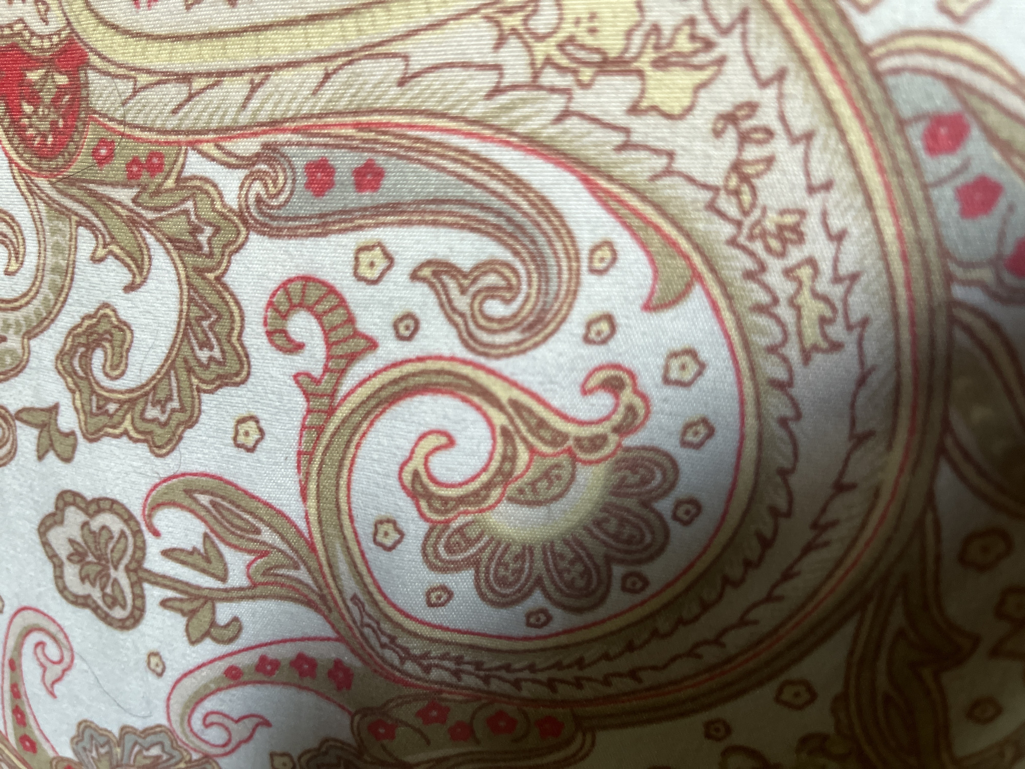 The image displays a close-up view of a fabric with a traditional paisley pattern. The pattern features swirling motifs in shades of gold, brown, and red against a light-colored background, possibly white or cream. Small floral accents in red are scattered throughout the design. The texture of the fabric is visible, showing a woven material. There is no identifiable location in the city of Orozbekov, Kyrgyzstan, as the image is solely focused on the textile design.