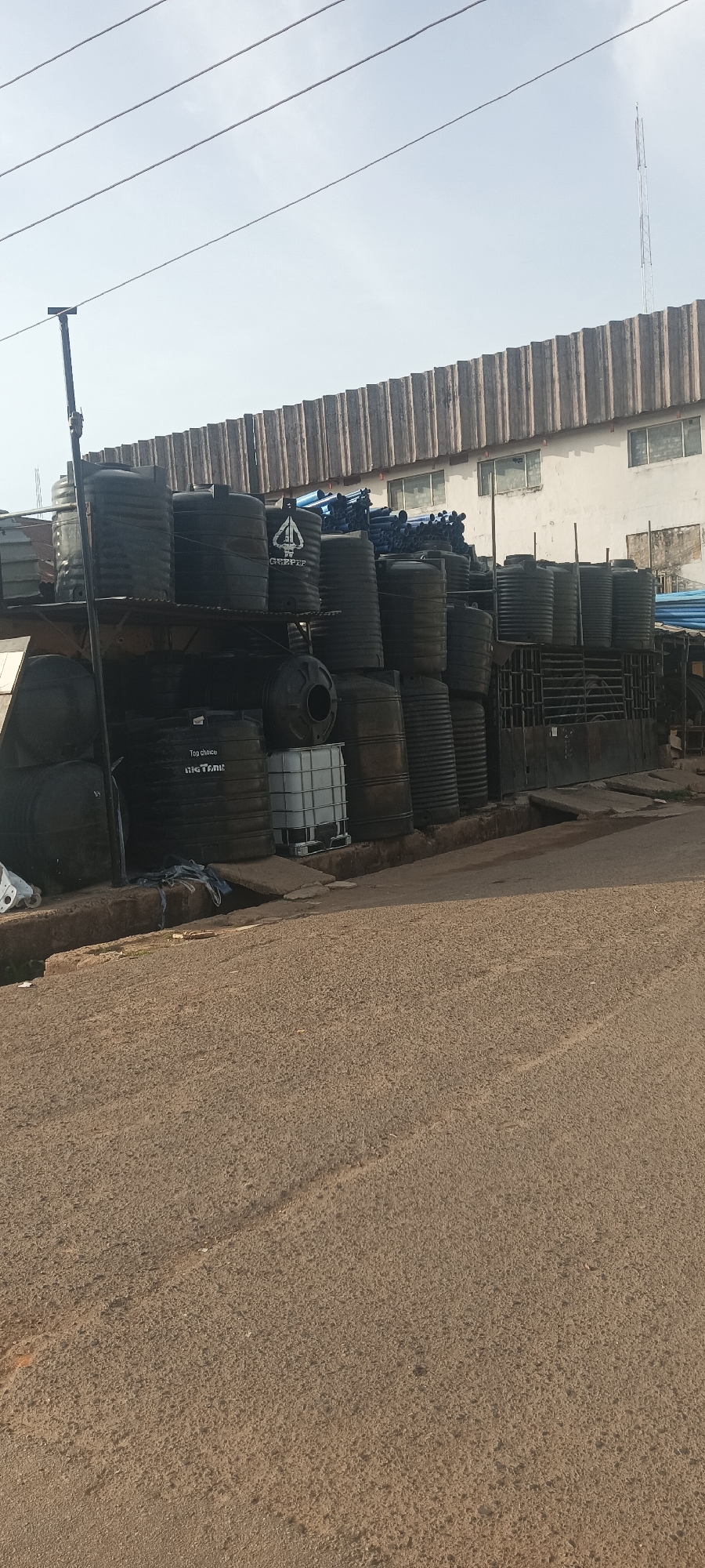 The image depicts a roadside scene in Jos, Nigeria, featuring a large accumulation of black plastic water storage tanks. The tanks are stacked on shelves beside a paved road. Some tanks have the branding "GRIZZ PET" and "BIG TANK" visible. Behind the tanks is a large building with a corrugated roof and windows. A utility pole is visible on the left side of the frame. The sky is overcast. The road surface is worn and displays signs of traffic.