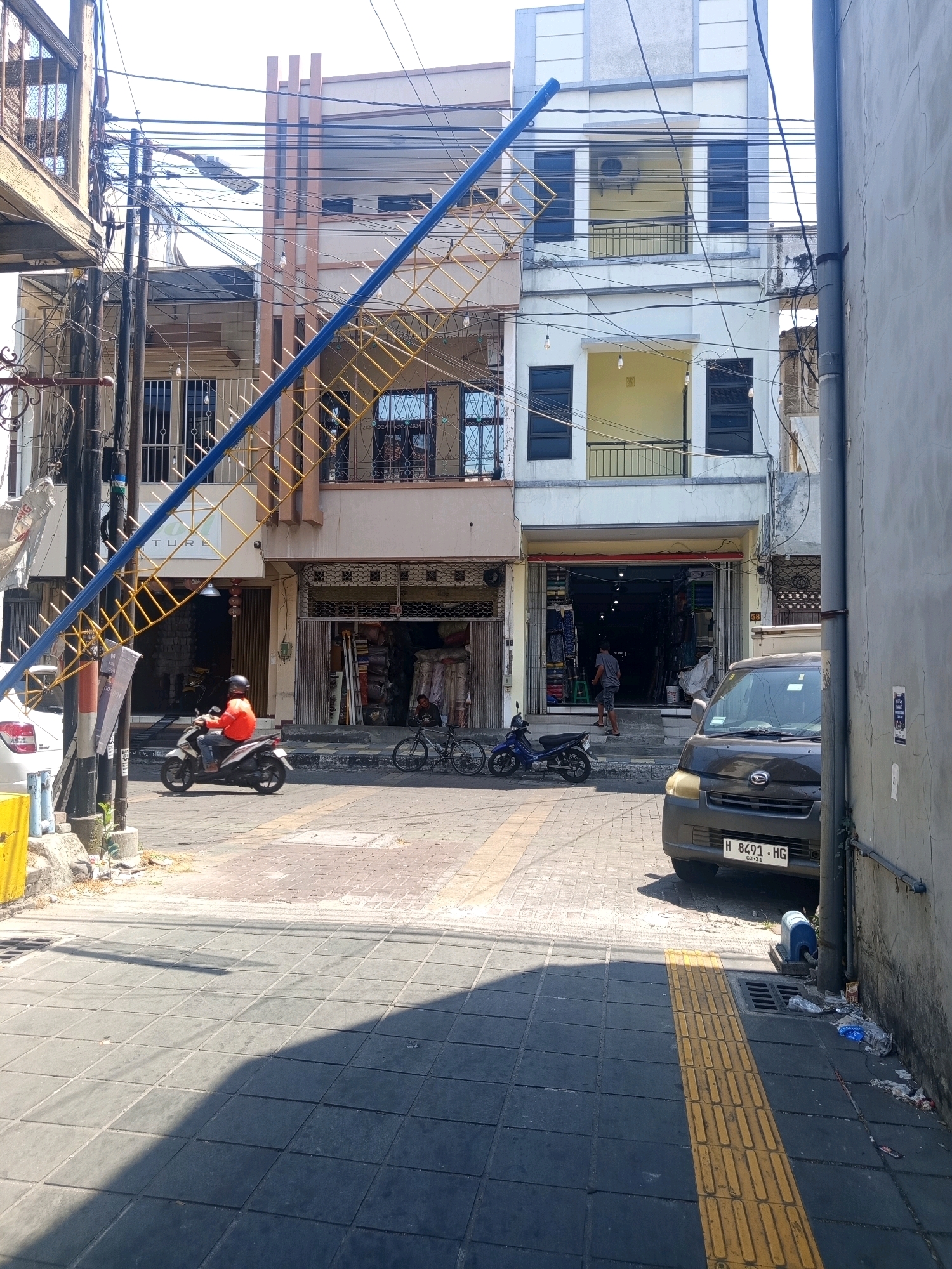 In the city of Semarang, Indonesia, on a sunny day, a street scene unfolds. A multi-story building with a light-colored facade and dark-framed windows dominates the right side of the image, its exterior adorned with a complex network of overhead electrical wires. To its left, a similar building, painted in a neutral tone, features balconies and windows, also draped with electrical wires. A striking diagonal element, a blue and yellow metal structure resembling an external staircase or support, cuts across the frame from the upper left towards the center.
On the street level, two shops are visible. The one on the left has its entrance partially obscured by a white car. The shop on the right has its facade open, revealing displays of goods, including rolls of fabric or carpet. A person stands in the doorway, while another is inside the shop.
The street itself is paved with interlocking bricks, with a yellow tactile paving strip running along the sidewalk on the right. A dark-colored van with a license plate reading "H 8491 HG" is parked on the right side of the street. In the foreground, a man wearing an orange jacket and a helmet rides a white motorcycle. To his right, a bicycle stands near the shop entrance. Further down the street, a blue motorcycle is parked. The environment appears to be an urban commercial area.