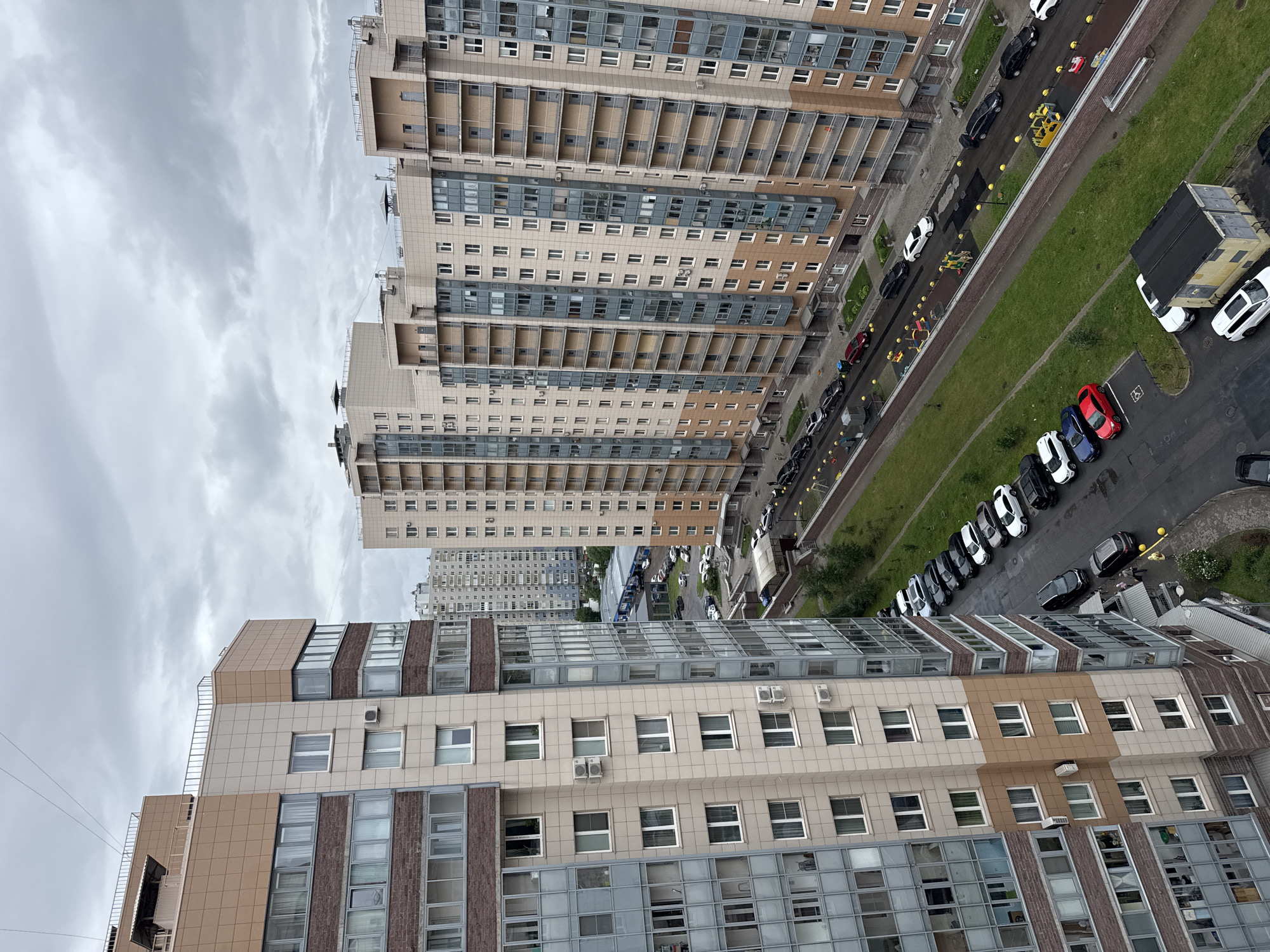 This media file shows an aerial view of a residential area with tall buildings and a street lined with parked cars. The scene depicts a cityscape under an overcast sky, suggesting a cloudy or possibly rainy day.
The primary subjects are the multi-story apartment buildings, characterized by their modern architecture with a mix of light-colored facades, glass balconies, and visible window units. The buildings are arranged in rows, with one building dominating the foreground with its light brown and grey exterior. Further in the distance, more buildings of similar design are visible, creating a dense urban environment.
Along the street running alongside the buildings, numerous cars are parked. These include a variety of colors and models, such as black, white, red, and blue vehicles. A few red and yellow toy vehicles are also scattered near a playground area. The street is bordered by a green grassy area, and in some parts, there are trees and bushes. A large dumpster or waste container is also visible on the side of the road.
There is no visible text in the image. The activity or event taking place appears to be a typical day in a residential urban setting, with no specific event or action prominently featured. The time of day is unclear due to the overcast conditions, but it seems to be daytime. The overall mood is neutral, reflecting a mundane urban scene. The presence of multiple identical apartment blocks suggests a planned residential development.