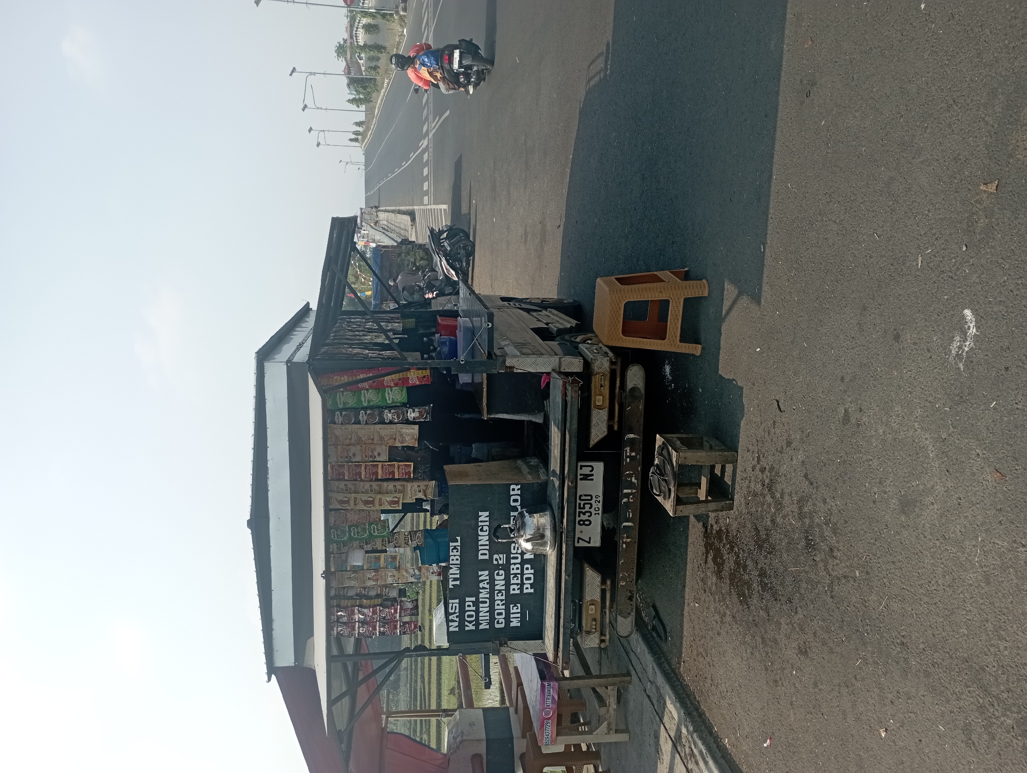 The image depicts a roadside food stall in Tasikmalaya, Indonesia. The stall is set up on the back of a small truck, which is parked on the side of a road. A variety of packaged food and drink items are displayed hanging from the stall's canopy. A sign on the stall lists "NASI TIMBEL," "KOPI," "MINUMAN DINGIN," "GORENG-2," "MIE REBUS," "LULUR POP," and "M." A metal kettle sits on the counter.
In the foreground, there are two small stools, one made of wood and the other of plastic. A pair of flip-flops rests on the wooden stool. The road surface is asphalt, with some scattered debris.
In the background, a motorcycle rider is seen on the road, heading away from the stall. The road stretches into the distance and appears to be an elevated highway or overpass. Streetlights are visible along the road. Beyond the road, there are some buildings and trees. To the left of the stall, a glimpse of a rice paddy field can be seen. The sky is clear and bright.