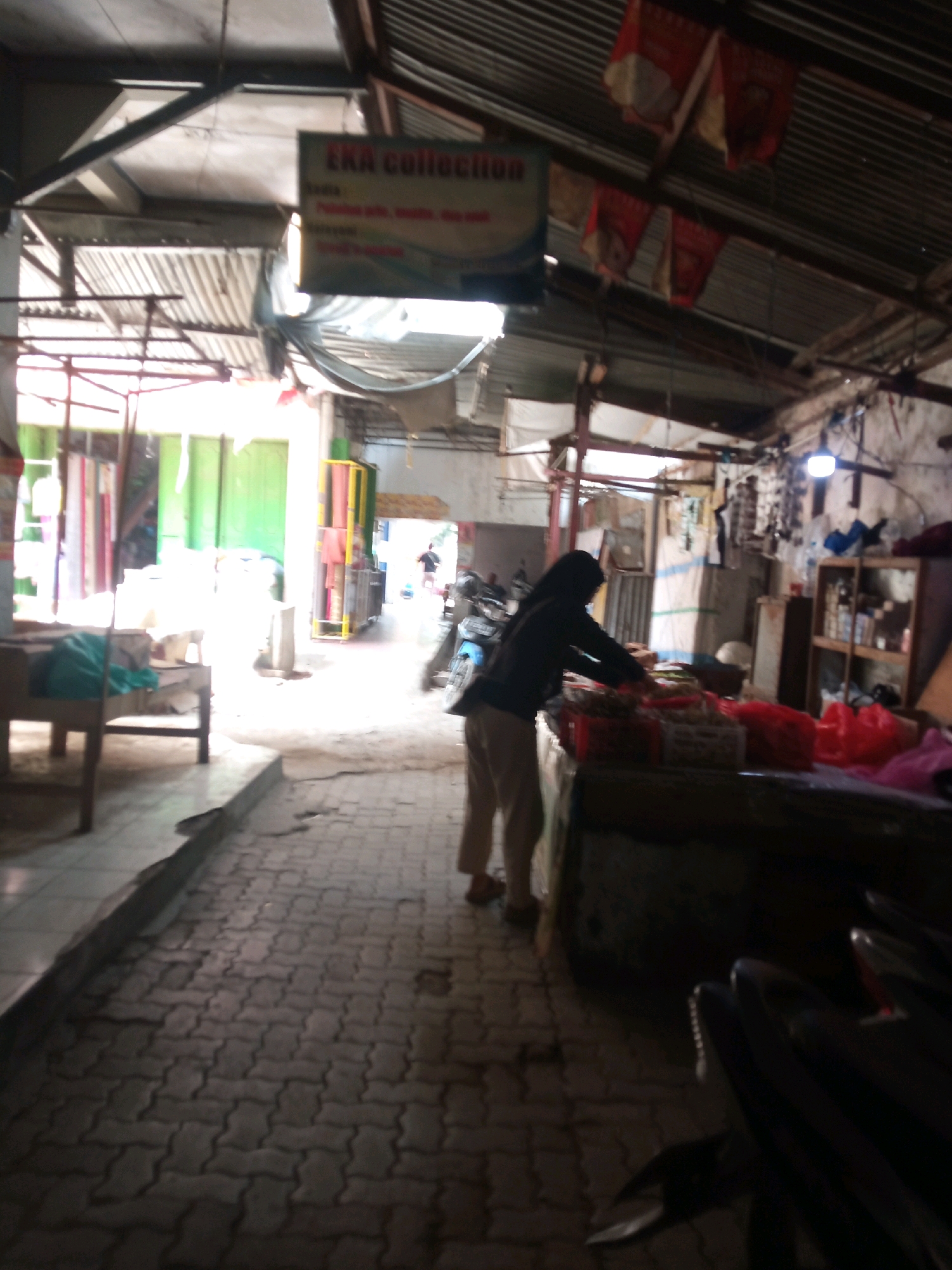 The image captures an outdoor market scene in Sambong, Indonesia. A woman, dressed in a dark hijab and light-colored pants, stands behind a stall displaying goods in red crates and bags, and possibly some fabric. She appears to be arranging or inspecting the items. In the foreground, paving stones form the ground surface. To the left, a wooden table with a blue cloth draped over it is visible. In the background, there are stalls with colorful fabrics or textiles, metal shelving units filled with items, and what appears to be a roof made of corrugated metal. A motorbike is parked to the right of the frame. The overall lighting suggests it is daytime, possibly overcast or with the sun filtered through the market structures. The scene depicts a typical marketplace environment in Indonesia, with goods displayed for sale and active commerce taking place.