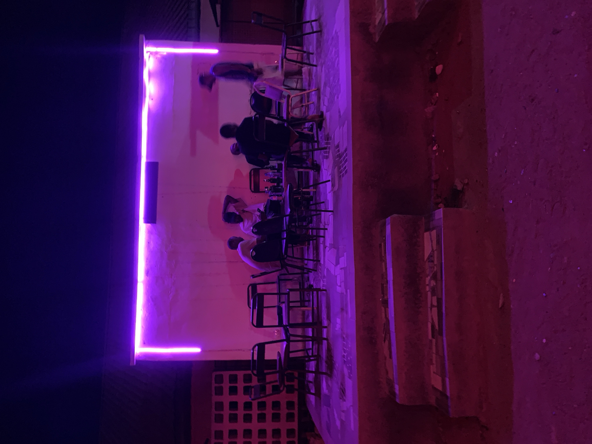 The image depicts an outdoor social gathering at night, likely a small bar or cafe in the vicinity of New Stadium, Nigeria. Several people are seated around tables, engaged in conversation. Bottles and drinks are visible on the tables. The area is illuminated by purple neon lights that frame a wall, casting a strong purple hue over the scene. The ground appears to be a combination of concrete and dirt. Chairs of metal and wood construction are scattered around. The darkness surrounding the illuminated area suggests a nighttime setting. A breeze block wall is visible in the foreground.