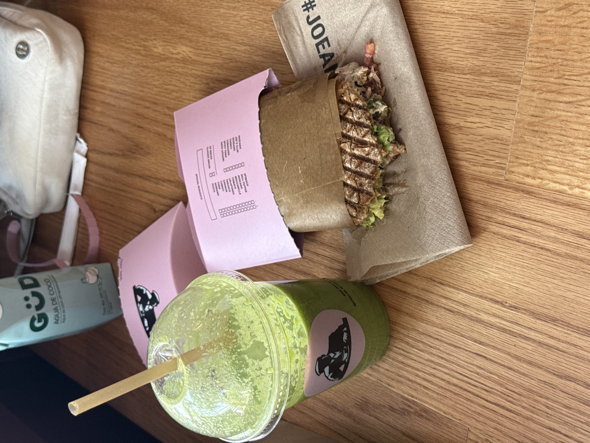 This overhead shot depicts a casual meal arranged on a light wooden table, likely in a cafe or restaurant setting in Mexico City, Mexico. The main subjects are a partially consumed grilled sandwich or panini, a green smoothie, and a carton of coconut water.
The sandwich is wrapped in brown paper and rests within a pink paper sleeve that serves as a menu or order form. Its grilled marks are visible, and the filling appears to include greens and other ingredients. Beside it, a clear plastic cup holds a vibrant green smoothie, adorned with a green label featuring a logo of a muscular male figure, and a beige straw. A light blue/green carton of "AGUA DE COCO" (coconut water) by the brand "GÜD" is also present, alongside a light-colored bag, possibly a Lululemon Everywhere Belt Bag, in the upper left corner.
The visible text on the sandwich wrapping includes "#JOEAN", likely part of "#JOEANDTHEJUICE". The pink sleeve lists sandwich options and ingredients such as "AVOCADO", "JOE'S CLUB", "TURKEY", "SPICY TUNA (VEGAN)", "GF BREAD", "CLASSIC BREAD", and various add-ons like "SPINACH", "MOZZARELLA", "JALAPEÑOS", and "PESTO". The smoothie cup has "RECYCLE HE" printed on it, suggesting a recycling instruction. The "GÜD AGUA DE COCO" carton is prominently labeled. The scene suggests a relaxed meal or snack enjoyed indoors.