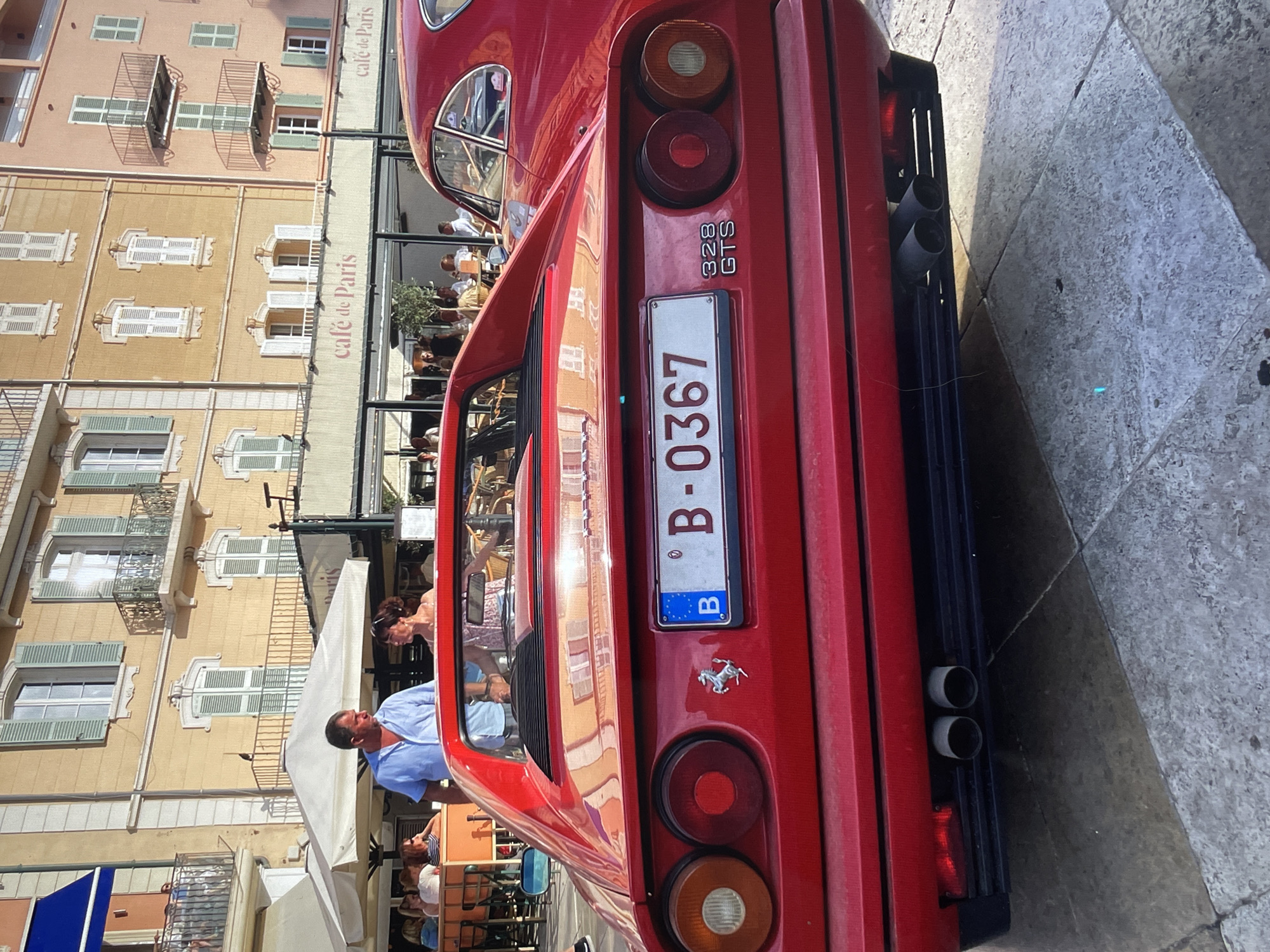Here's a detailed description of the image:
**Overall Scene:** The image is taken outdoors in a city, likely a tourist area, due to the presence of a "Café de Paris" sign and architectural style. The primary subject is the rear of a red Ferrari 328 GTS, with a Belgian license plate reading "B-0367".
**Environment:** The Ferrari is parked on a stone-paved surface. Behind the car, buildings with multiple floors and balconies, featuring a Mediterranean architectural style, are visible. A "Café de Paris" sign is also visible, indicating a cafe or restaurant in the vicinity.
**People:** Several people are visible in the background, likely patrons of the cafe. A man in a light blue shirt appears to be near the car.
**Objects:** Besides the car and buildings, elements like street lamps, awnings, and cafe tables are seen, contributing to the urban setting.
**Car Details:** The Ferrari's rear lights, exhaust pipes, and the iconic Ferrari emblem (prancing horse) are clearly visible. The "328 GTS" model name is also visible on the right side. The Belgian license plate is prominently displayed.