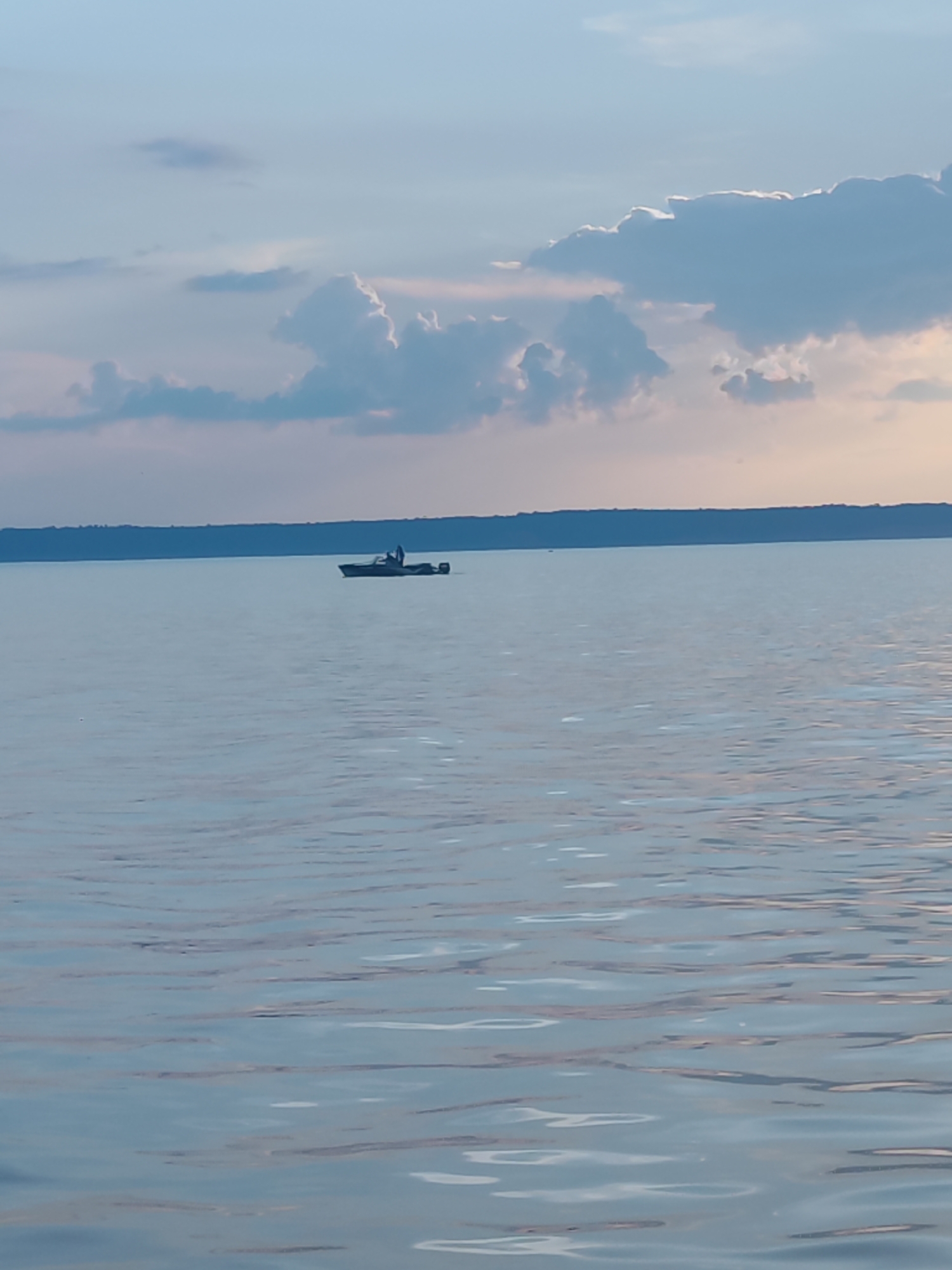 A boat with a person on board is seen in the distance on a large body of water under a cloudy sky. The boat is dark-colored and appears to be a motorboat, with a single person standing in it. The water is calm with gentle ripples, reflecting the light blue and white of the sky and clouds. A dark, tree-lined horizon is visible in the background. The overall scene is serene and suggests a quiet day on the water. This scene could be located in the city of Галево, Russia, given the presence of a large body of water.