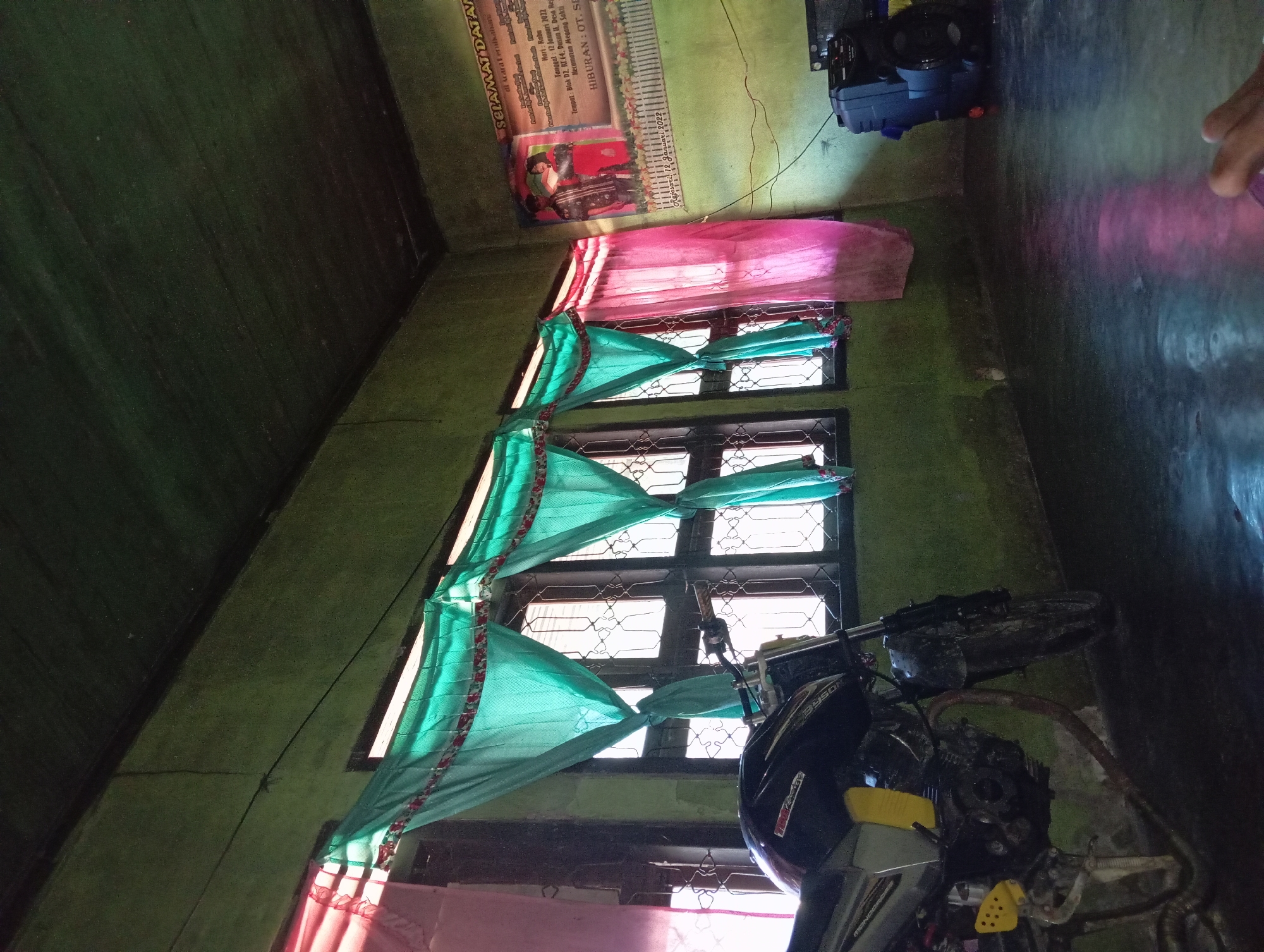 The image captures an indoor scene, likely a room in a residence, in the city of Rejosari, Indonesia. A motorcycle, predominantly black with yellow accents on the side, is parked indoors to the left of the frame. In the background, multiple windows are visible, draped with colorful curtains – teal and pink – that diffuse the natural light entering the room. A poster with text in Indonesian, likely announcing an event, is mounted on the wall to the right, featuring images of two individuals. Adjacent to the poster is a large black speaker. The walls and ceiling appear to be painted a muted green. The floor is dark and reflective, showing some light patterns. The overall impression is a lived-in space, possibly a garage or living area, with personal belongings present.
