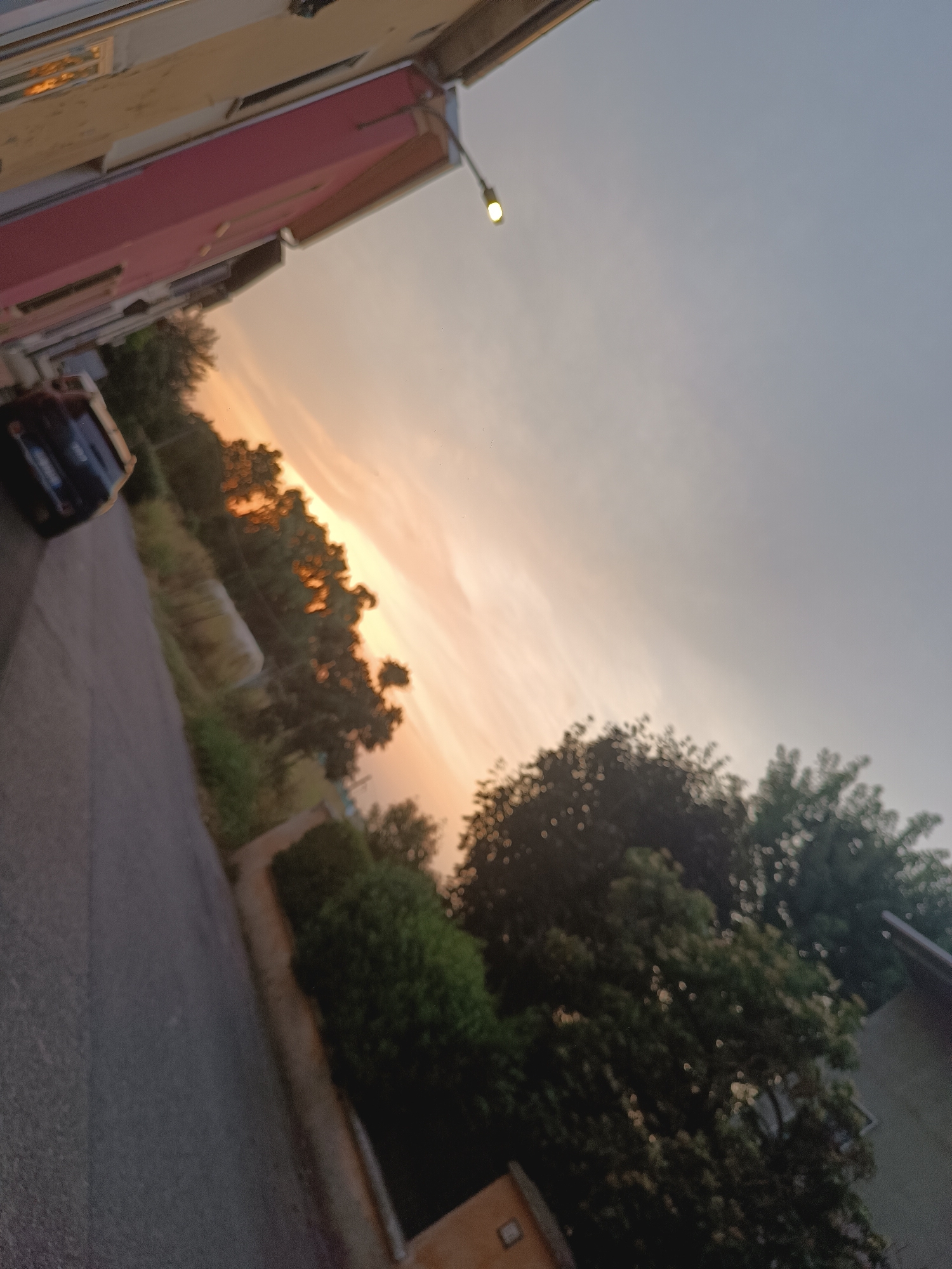 The image depicts a street scene in Viadana, Italy, likely during dusk or dawn, given the warm hues in the sky. The perspective is angled, giving a sense of movement or tilt. On the left, a dark-colored car is parked or driving slowly on an asphalt road. Adjacent to the road, a building with a pink facade and beige upper sections is visible. Further into the scene, lush green trees and vegetation line the roadside, their leaves softly blurred. The sky transitions from a soft orange and yellow near the horizon, suggesting the sun's position, to a pale blue-gray overhead. A single light fixture with a lit bulb is prominent in the upper left quadrant of the image, suspended against the sky. The overall impression is a quiet, residential street bathed in the soft light of twilight.