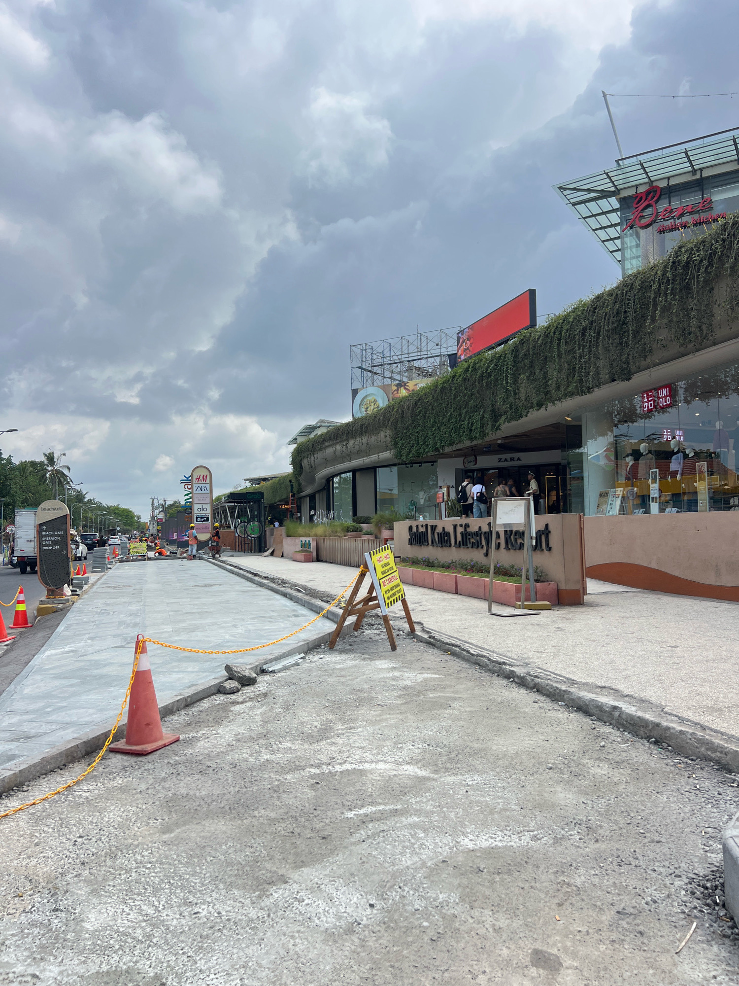 This image depicts a street scene in Kuta, Indonesia, with a focus on ongoing construction work along the sidewalk. In the foreground, a newly paved section of sidewalk is visible, bordered by a construction zone marked with orange cones and yellow caution tape. The ground is a mix of gravel and unfinished concrete, indicating active construction.
Further down the street, several shops are present, including a Zara store, and a sign for "Beachwalk Shopping Center." Across the street, a variety of vehicles are driving, and there are palm trees lining the road, typical of a tropical location. The sky is overcast with heavy clouds, suggesting a humid or potentially rainy day. Several construction workers can be seen on the street, some wearing safety vests and hard hats, engaged in their tasks. A prominent sign reads "Sahid Kuta Lifestyle Resort," identifying the location. The overall impression is one of a busy urban area undergoing development.
