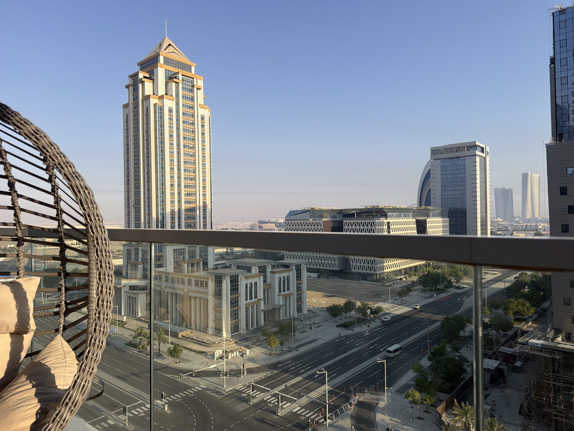 An aerial view captures a cityscape under a clear blue sky, with the foreground dominated by a wicker hanging chair with two beige cushions. The chair is positioned on a balcony, with a glass railing in front of it. Beyond the railing, a wide, multi-lane road curves through a modern urban environment.
Several buildings are visible, including a tall, slender skyscraper with a golden-toned facade and a stepped roofline. To the right of this skyscraper are several other contemporary buildings, some with curved architectural elements and others with patterned facades. In the distance, more skyscrapers rise against the horizon.
The road is marked with white lines, and several cars and a white bus are seen on it. Pedestrian crossings are visible, along with traffic lights and street lamps. The landscape is dotted with palm trees and other greenery, indicating a warm climate. Construction work is evident in some areas, with scaffolding and partially built structures. The overall impression is one of a modern, developing city.