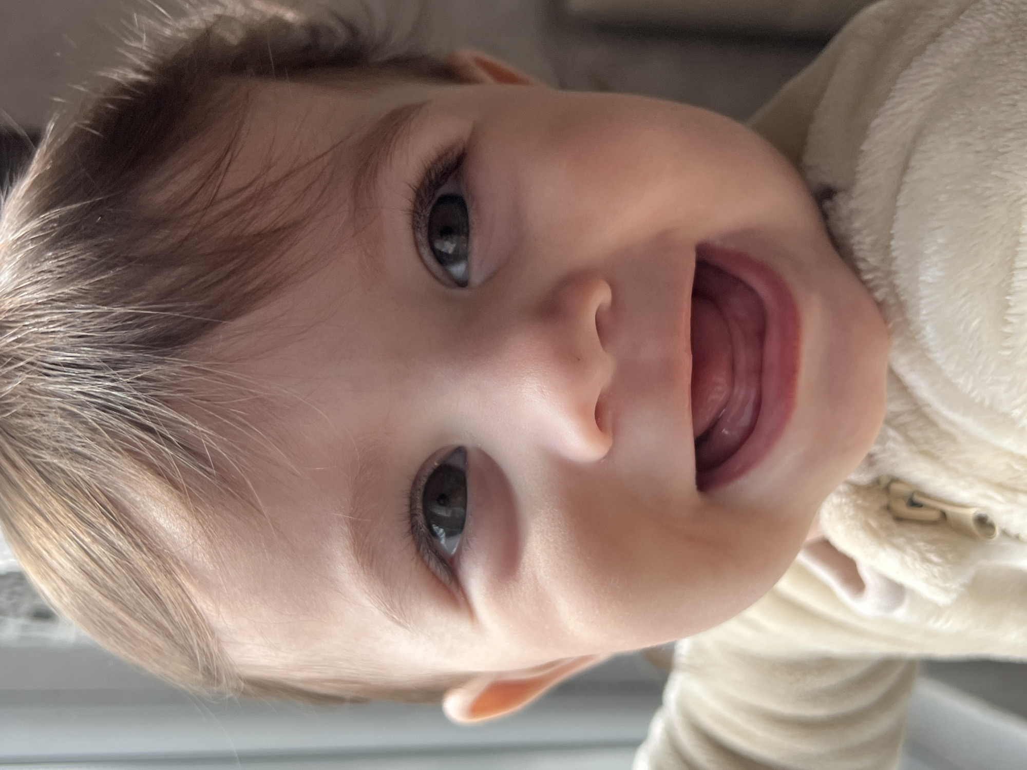 A close-up, frontal portrait captures an infant with fair skin, dark brown eyes, and light brown, wispy hair, styled with a fringe across the forehead. The infant is smiling broadly, mouth open, revealing the tongue but no teeth. The subject is wearing a cream-colored, fuzzy, long-sleeved garment, possibly a onesie or jacket, with a partial zipper visible on the upper chest. The background is indistinct and blurry, indicating an indoor setting. A strong natural light source, likely a window, is positioned to the infant's left, illuminating the left side of the face. The visual evidence does not provide any specific markers to identify a location within the city of Elena, Bulgaria.