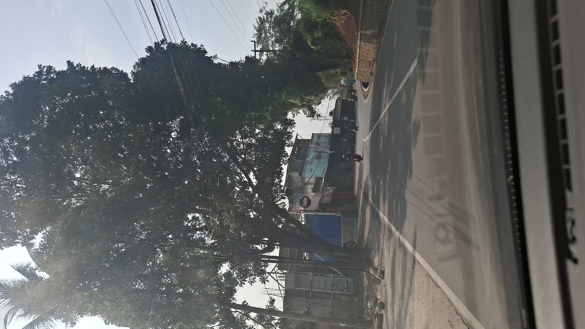 This image captures a view from a vehicle driving on a paved road in Cilaku, Indonesia. The road is lined with trees, and on the left side, there are several buildings, including structures with blue and white corrugated metal siding, and a sign with the word "TACO". A few motorcycles are visible on the road, with one rider in the foreground on the left and others further down the road. Overhead power lines run along the scene. The lighting suggests it is daytime, with sunlight filtering through the trees, casting shadows on the road. The environment appears to be a mix of residential and commercial structures with natural vegetation.