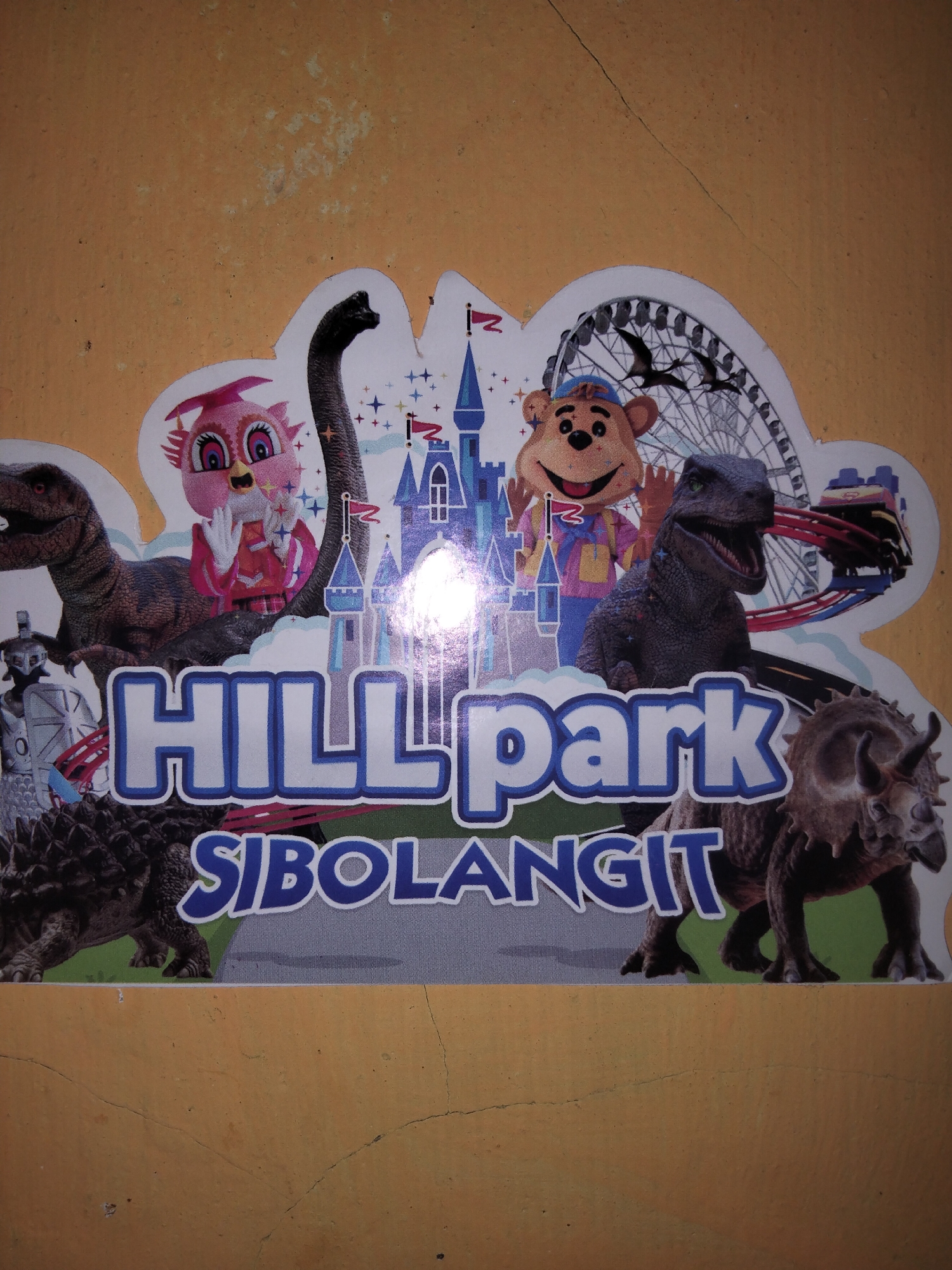 The image is a photograph of a colorful sticker or decal applied to a textured, orange-yellow wall. The sticker depicts various cartoon characters and illustrations, including dinosaurs, a stuffed bear, an owl character in a graduation cap, a Ferris wheel, a roller coaster, and a castle. The text "HILL park" is prominently displayed in large, stylized blue and white letters, with "SIBOLANGIT" written below it in similar lettering. The scene appears to be an advertisement or signage for a place called Hill Park Sibolangit, an amusement park located in the Karo Regency of North Sumatra, Indonesia, near the city of Medan and not Payagaboh. The lighting in the photo is uneven, with some bright spots suggesting artificial light sources, and there are visible cracks in the painted wall surface.