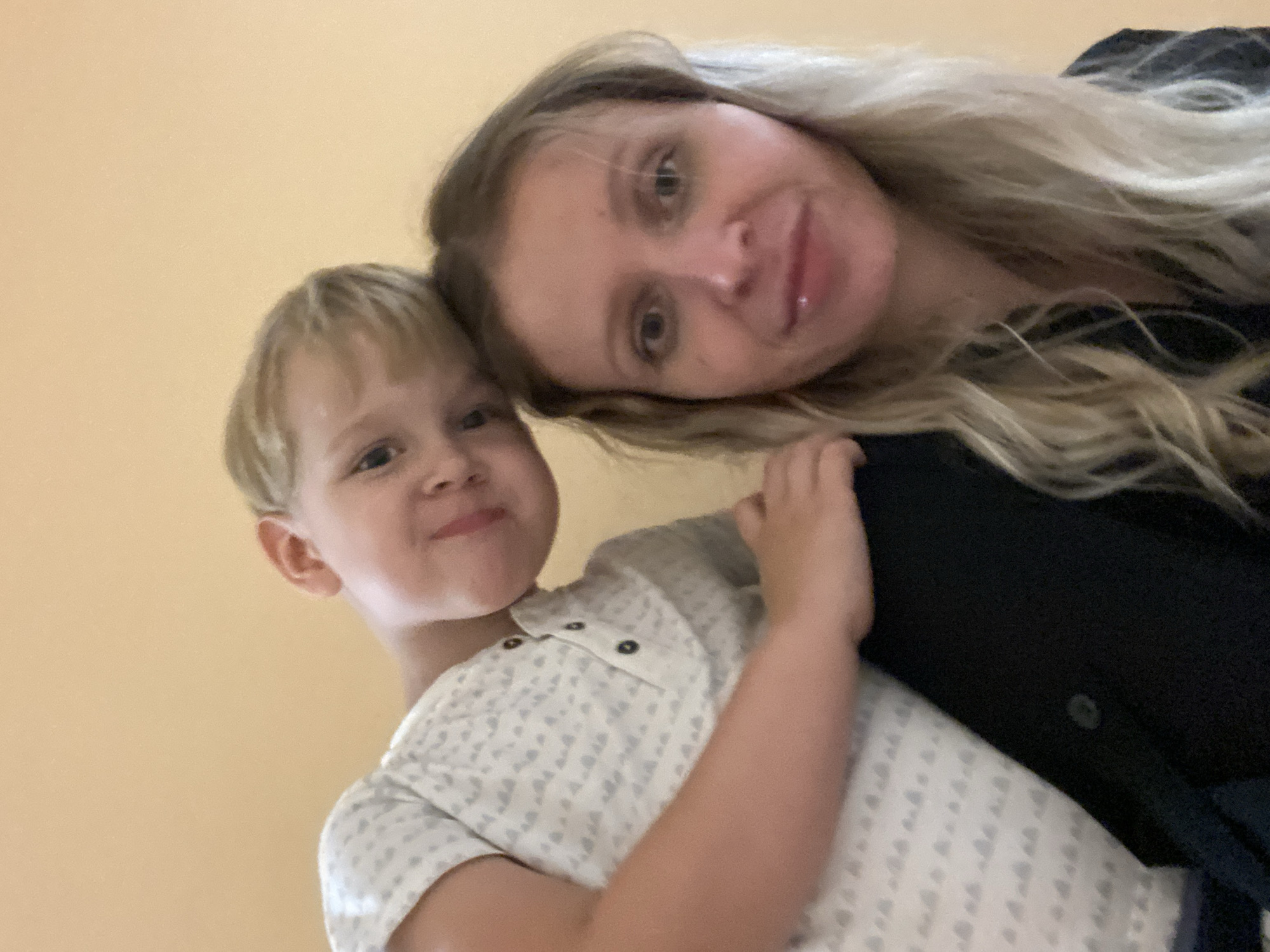 In this close-up, low-angle selfie, a young child with blond hair leans over the shoulder of an adult woman, resting his head on her head. Both individuals are looking towards the camera. The child wears a white t-shirt with a subtle grey pattern. The woman has blond hair and is wearing a dark top. The background is a plain, light orange wall, characteristic of an interior setting. The image quality is somewhat grainy and lacks sharp detail, suggesting it was taken with a mobile device. The overall impression is an informal, candid moment captured between a mother and her child. No specific details of Shiryaevo, Russia, are discernible from the provided image.