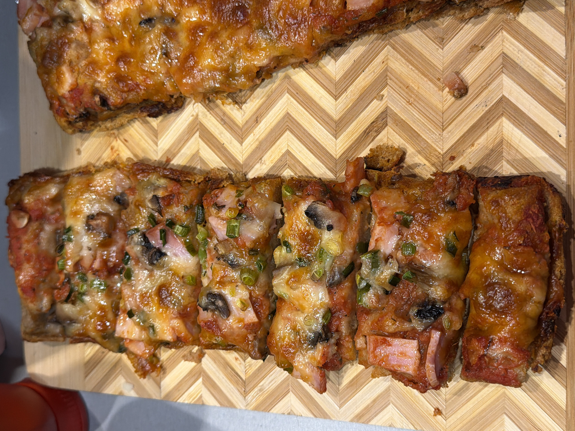 The image shows sliced pieces of cooked rectangular pizza on a wooden cutting board with a chevron pattern. The pizza appears to have a thick, toasted crust, tomato sauce, melted cheese, ham, mushrooms, and green onions. The pizza is cut into approximately six individual slices, with another piece resting above. The scene lacks any indication of location within Varna, Bulgaria or information about people.