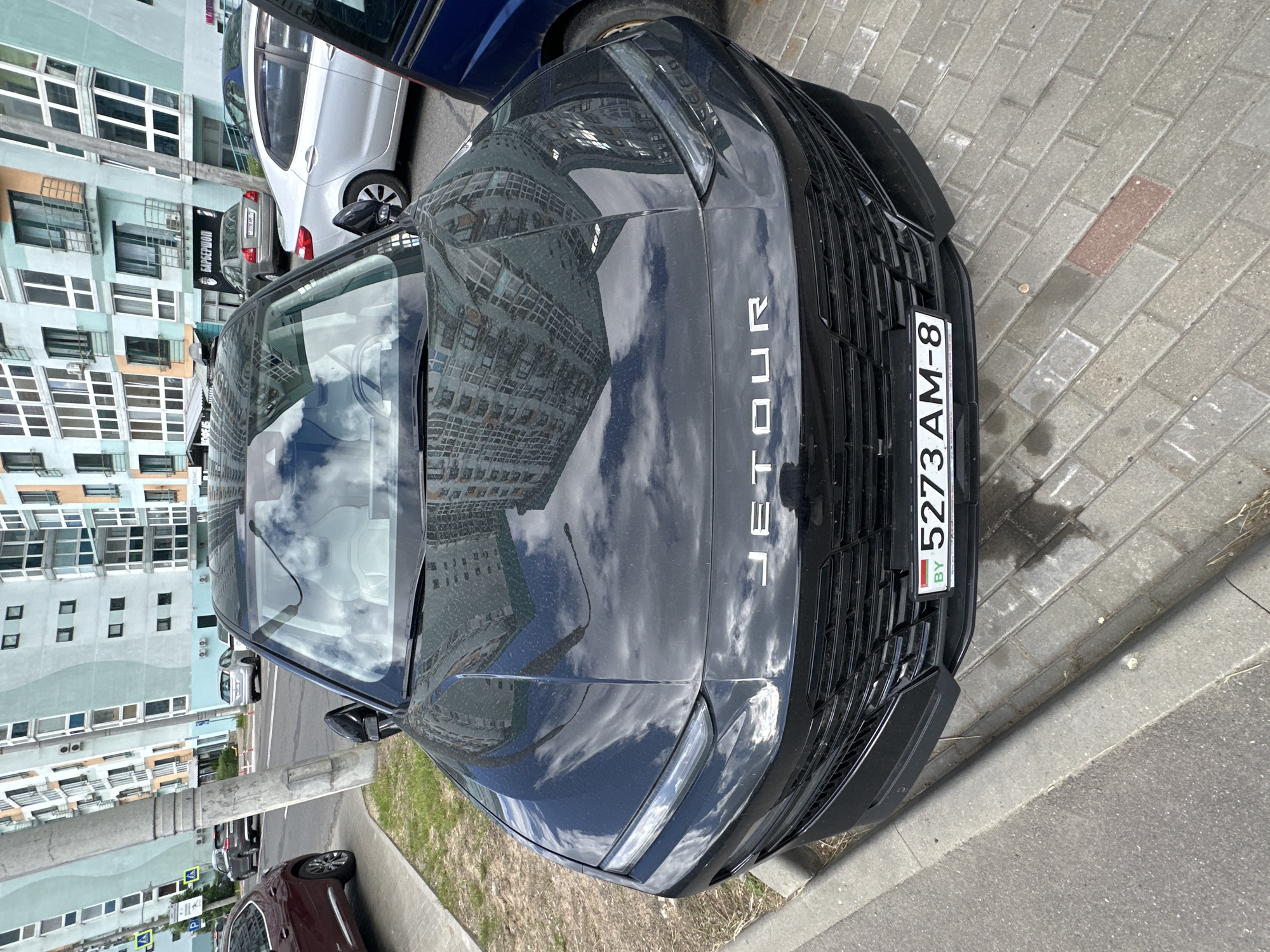 The image shows the front of a black Jetour car parked on a paved sidewalk. The car's license plate reads "5273 AM-8" and includes the "BY" country code, indicating it is registered in Belarus. The reflection on the car's hood shows a multistory building with many windows, suggesting an urban environment. Other cars are parked nearby, with one silver car and part of a red car visible. The backdrop features a modern building with a blue and white facade. The scene appears to be in Minsk, Belarus, based on the car's license plate.