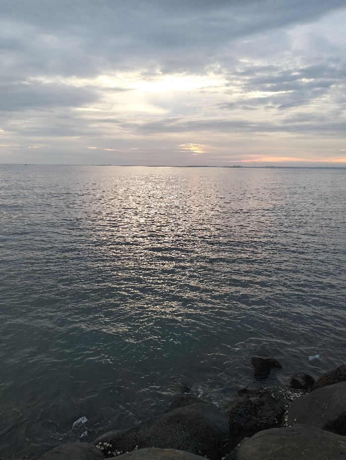 A seascape with calm, rippled water reflecting sunlight under a cloudy sky. The horizon line is visible with a faint outline of land. Large, dark rocks are present in the lower right foreground. The sky has various shades of gray and white, with brighter patches where the sun's light penetrates.