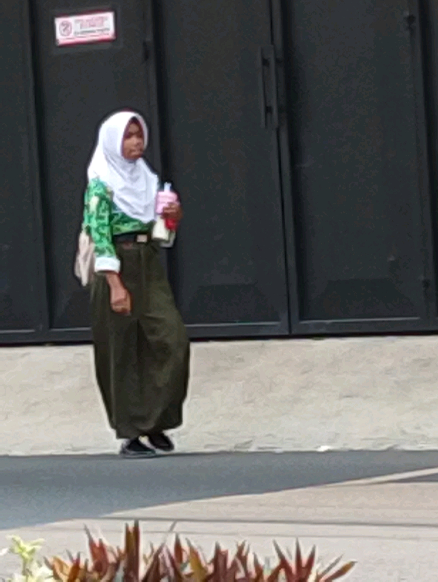 A young woman wearing a white hijab and a green patterned long-sleeved shirt, a dark olive green skirt, and dark sneakers is standing in front of a dark gray industrial-style gate. She is holding a pink and white bottle in her right hand. A white shoulder bag is slung over her left shoulder. The ground in front of her is paved with a light-colored, rough textured material, and a darker gray asphalt road is visible in the foreground, with some reddish-brown and green plants. A sign with a red border and white text is affixed to the gate above and to the left of the woman. The scene appears to be in an outdoor urban or industrial area.