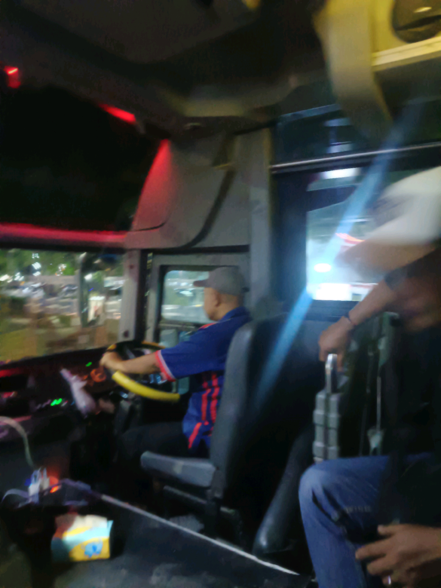 The image shows the interior of a bus at night, likely in Kedungrejo, Indonesia. A man wearing a grey cap and a blue shirt with red stripes is seated in the driver's seat, gripping the yellow steering wheel. His right hand is on the wheel, and his left hand is not visible. The dashboard is dimly lit with green and red lights. Outside the bus, through the windshield, streetlights and blurry car lights indicate a city environment. To the right of the driver, another person's arm and shoulder are visible, wearing a dark shirt and blue jeans. This person appears to be a passenger, and their hand is resting on a lever. A roll of tissues with a yellow and blue design is placed on the floor in front of the driver's seat. The overall lighting is dark with artificial lights from within the bus and outside contributing to the scene.