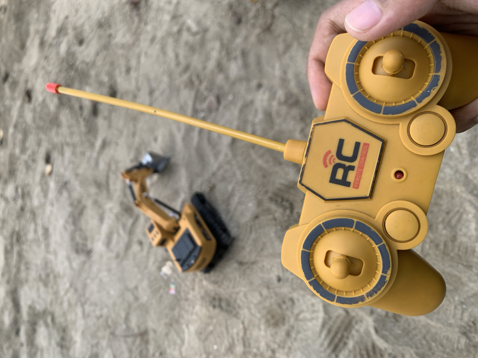 The image depicts a yellow RC excavator toy on a sandy surface in Tanjung Limau, Indonesia. A yellow remote control with two joysticks is held by a person's hand. The antenna of the remote is extended. The RC excavator toy is located on the left side of the image.
