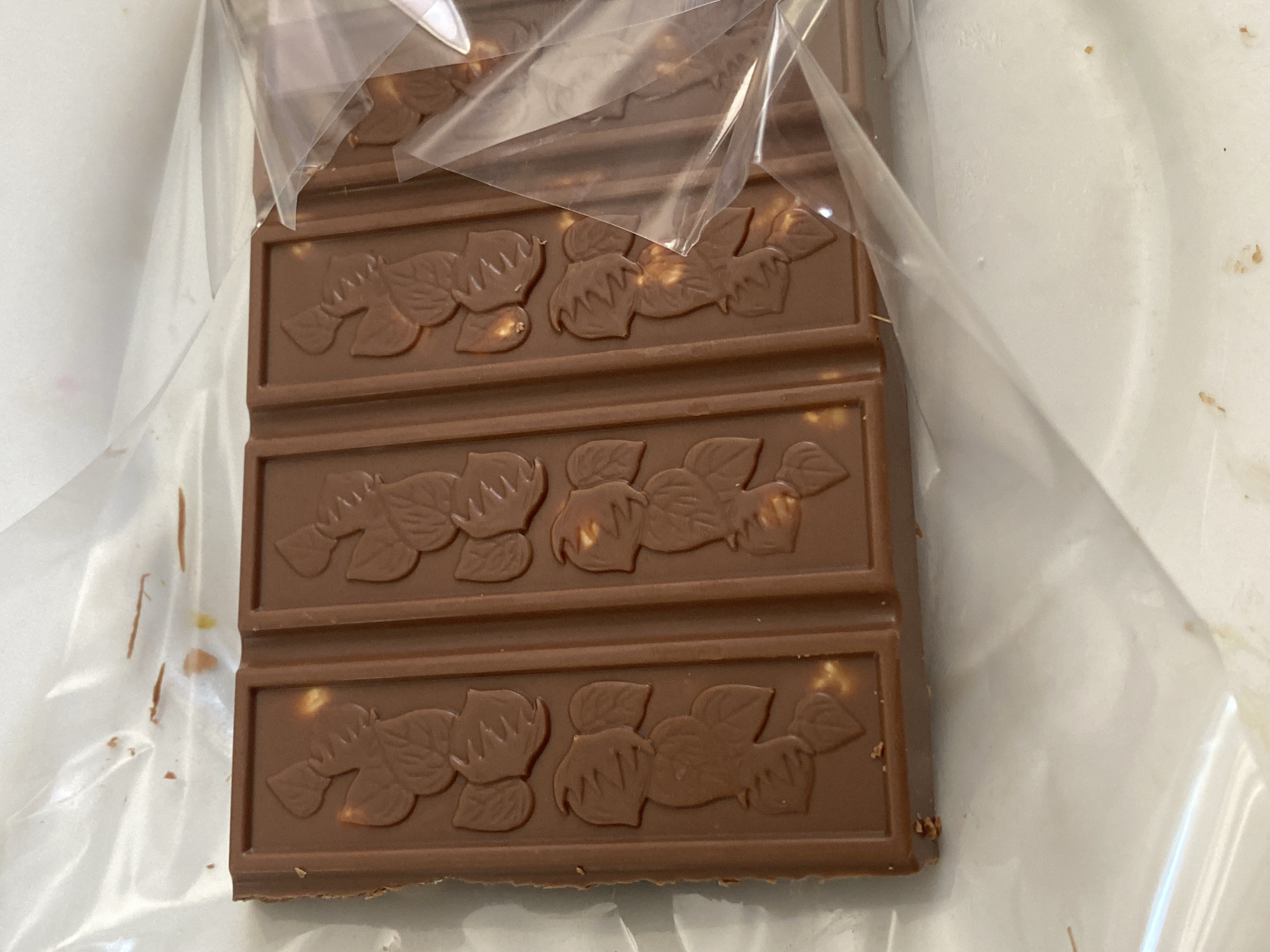 A close-up, overhead shot shows a broken chocolate bar, with several squares missing, lying on a white surface, possibly a table or counter, partially covered by a clear plastic wrapper. The chocolate bar has raised patterns of leaves and nuts embossed on each segment. The wrapper has some smudges and possibly food debris on it. The background is out of focus, but appears to be a light-colored surface with some scattered brown specks. There is no indication of the city of Chornomorsk, Ukraine in the image.