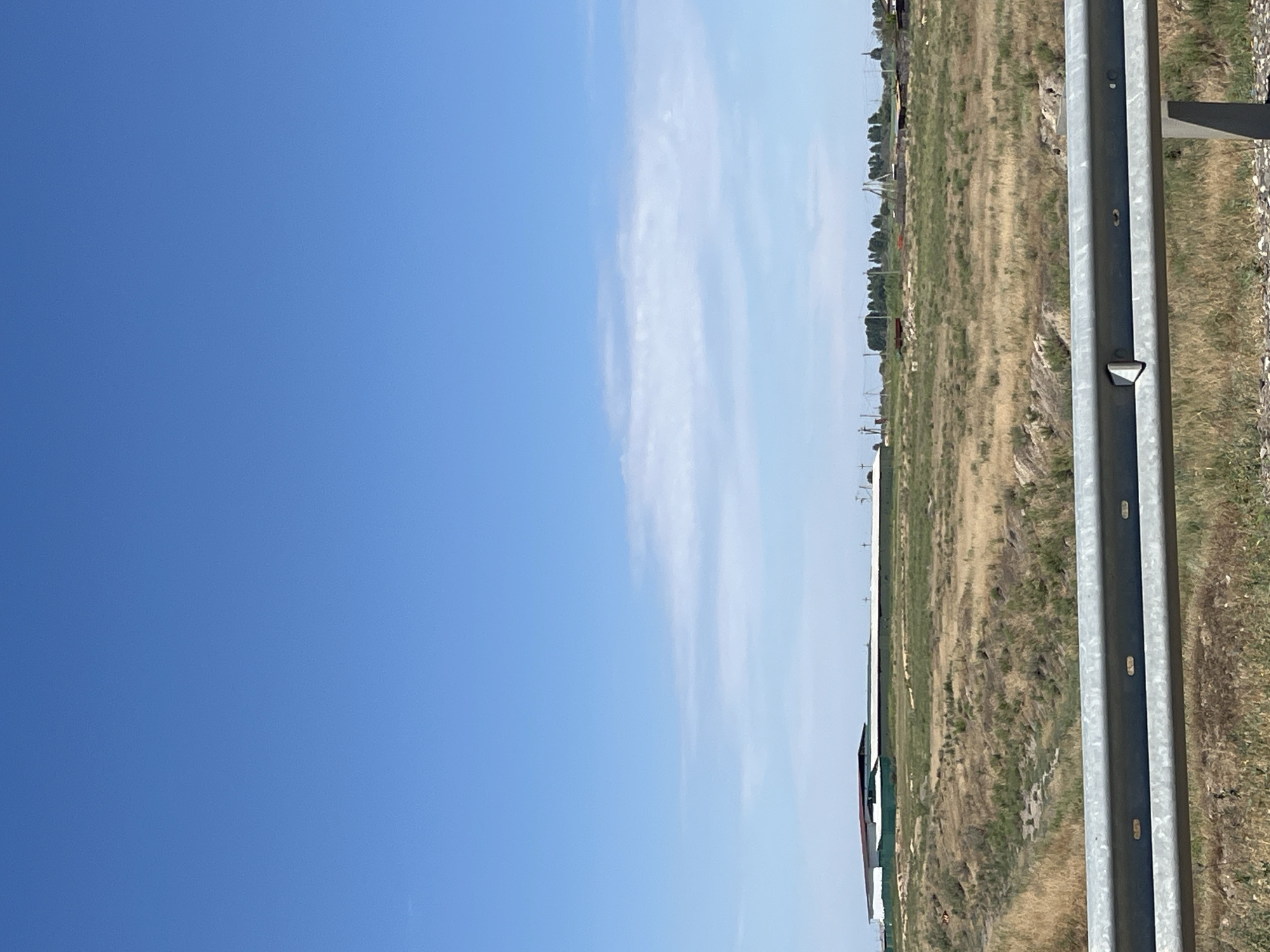 The image shows a wide, clear blue sky over a rural landscape. In the mid-ground, there are agricultural or industrial buildings with white and red roofs, surrounded by green fencing. The foreground features a dry, grassy field with some scrub vegetation and a metal guardrail running horizontally across the bottom of the frame. In the far distance, a line of trees and some structures suggest a settlement. The scene appears to be an outdoor view, possibly from a roadside, in a dry, sunny climate. There are no people or animals visible in the image. The location is identified as Bakhtemir, Russia.