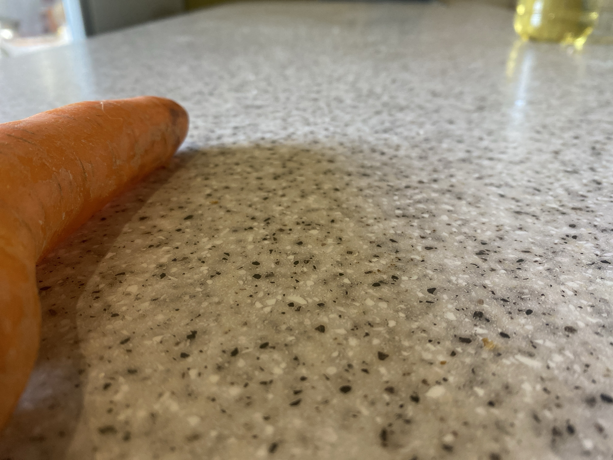 The image shows a close-up, low-angle shot of the end of a carrot resting on a speckled countertop. The carrot is a vibrant orange and appears to be fresh. The countertop is a light gray with darker speckles, giving it a textured appearance. The lighting suggests an indoor setting, possibly a kitchen, with a soft light casting a shadow from the carrot onto the countertop. In the upper right background, a blurred yellow liquid can be seen in a container, possibly oil or juice. The focus is on the carrot and the texture of the countertop, with the background elements out of focus. No people are visible, and the overall impression is a simple still life of food preparation ingredients. The scene is static, with no activity taking place. There is no visible text in the image.