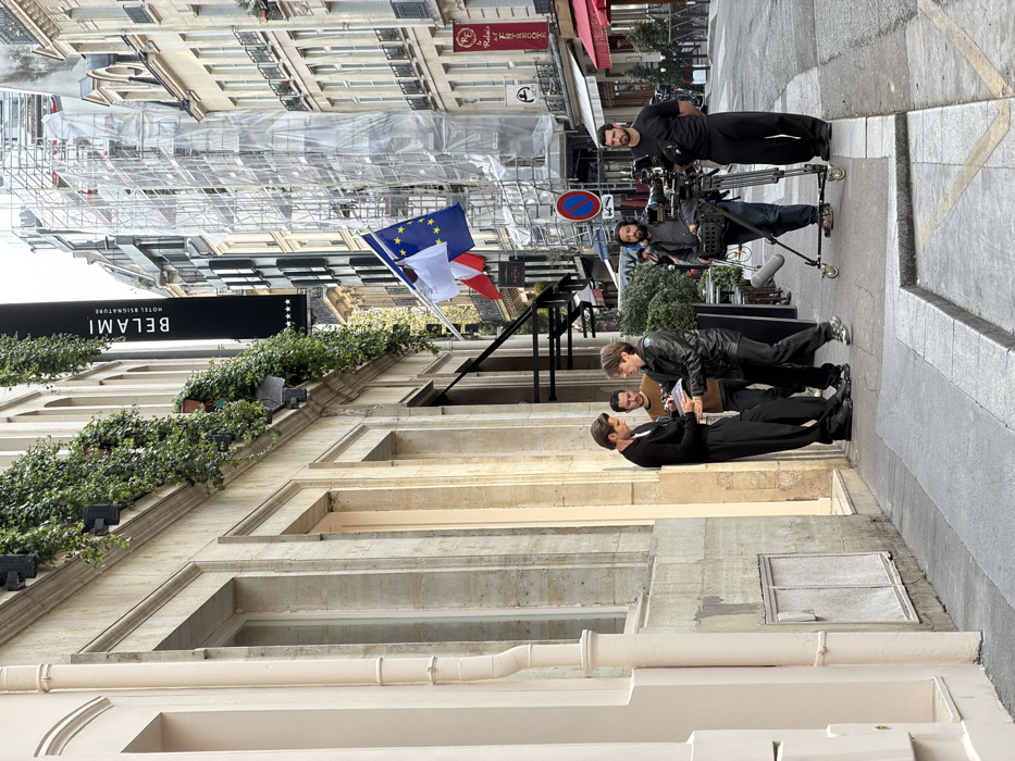 An outdoor scene depicts a film or photoshoot production taking place on a paved street in Paris, France, identifiable by the "BELAMI HOTEL & SIGNATURE" sign at 7-11 Rue Saint-Benoît.
On the left, a tall man with dark hair, wearing a black cardigan over a white shirt and black trousers, leans against the light-colored stone facade of the Hôtel Bel Ami. He is holding a small book or document in his left hand and appears to be signing it with his right hand. Facing him is a man in a black leather jacket and dark trousers, holding the item for signing. Behind the man in the leather jacket stands a man with a beard, wearing a brown long-sleeved shirt, observing the interaction.
To the right of this group, two crew members are present. One man with a beard, dressed in a dark shirt and jeans, operates a professional video camera mounted on a wheeled tripod (dolly) with a "dead cat" microphone attached. Standing to his right is another man in a black t-shirt and black trousers, with his hands clasped behind his back.
The street features a raised sidewalk with a curb. A blue circular "No Parking/No Stopping" sign with a red diagonal line is visible on a pole near the camera equipment. Overhead, a French tricolor flag and a European Union flag are displayed from an awning structure. The Hôtel Bel Ami facade is adorned with ivy in window boxes or ledges and features a black vertical sign with three white stars. In the background, other multi-story Parisian buildings line the street, with one structure extensively covered in scaffolding and white sheeting. Red awnings of other businesses are also visible further down the street. The sky appears overcast.