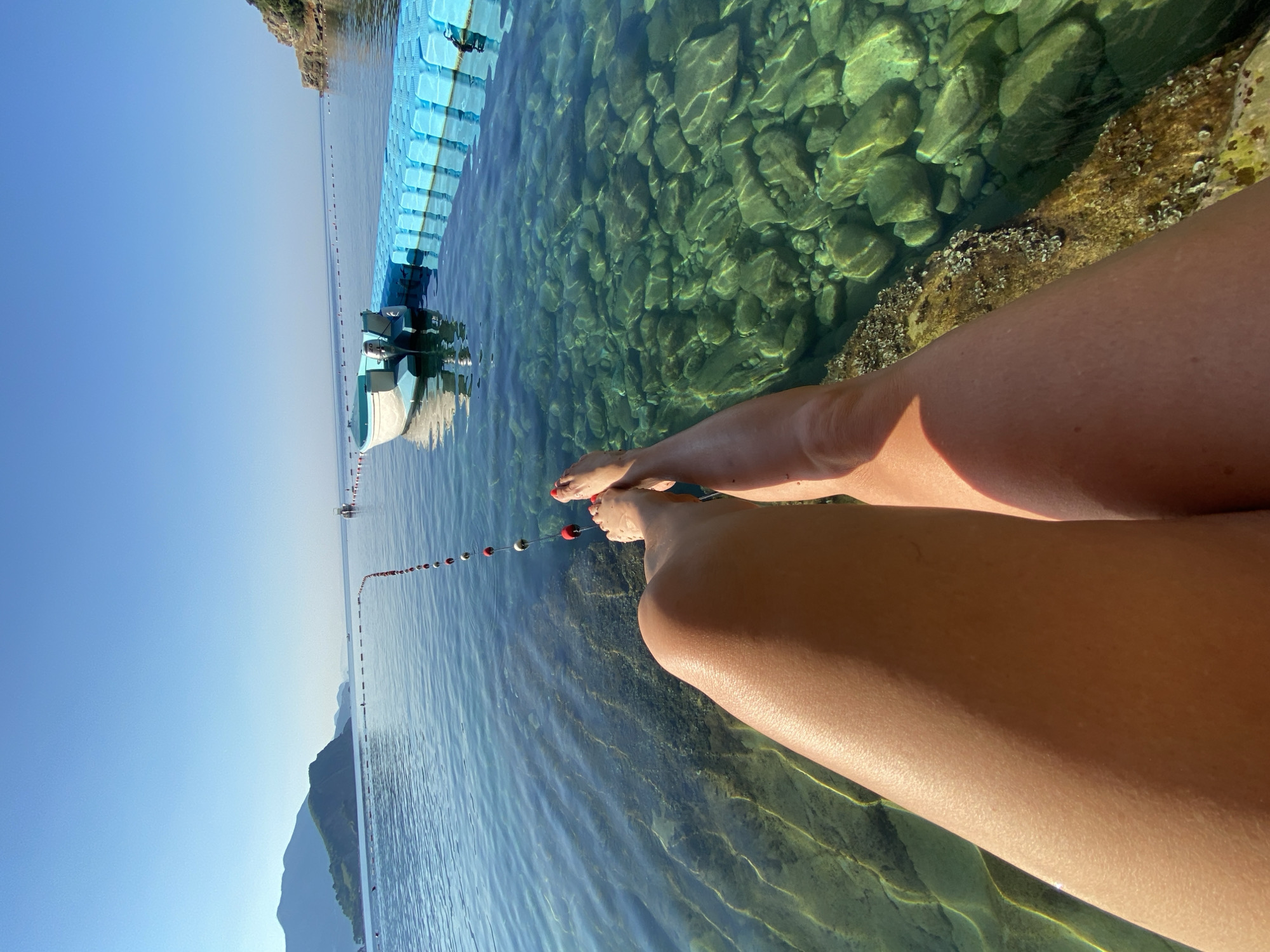 A person's legs are visible from the knee down, resting on the edge of a rocky shoreline. The water is crystal clear, revealing the rocky seabed below. In the background, a white boat with a blue trim is docked next to a blue floating platform. The sea stretches out to the horizon, with distant mountains under a clear blue sky. A line of buoys marks an area in the water. The scene appears to be located in Budva, Montenegro, possibly in the vicinity of a beach or marina. The lighting suggests it is a sunny day, likely in the morning or late afternoon.