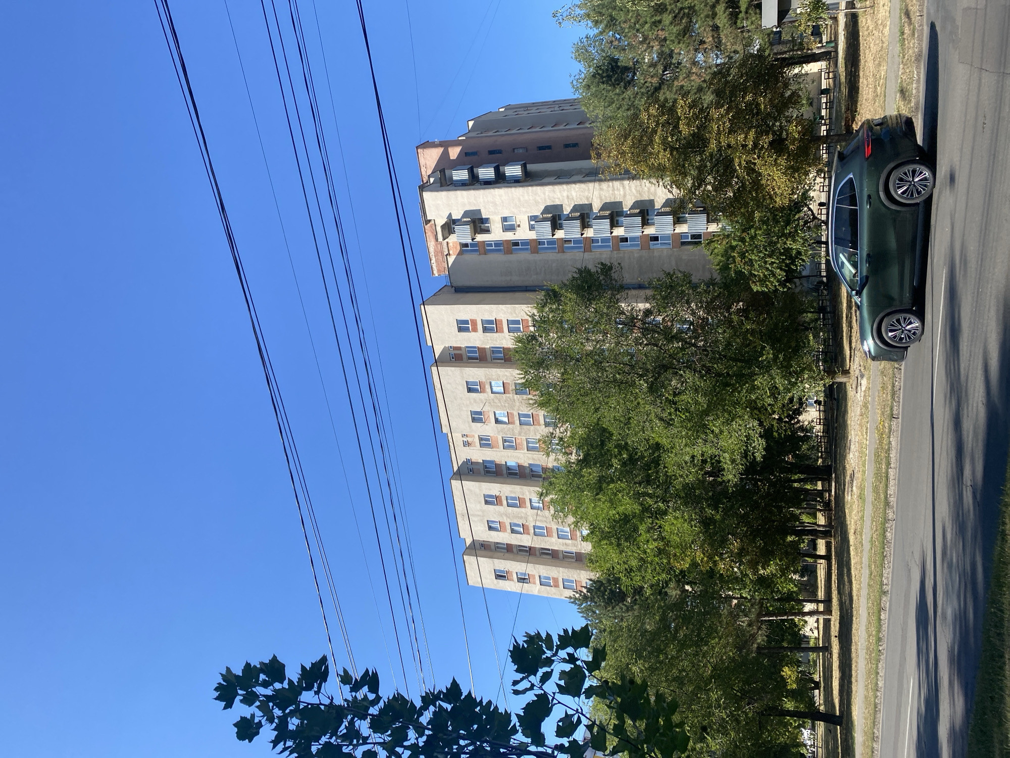 This is a vertical, medium-shot, eye-level, outdoor photograph taken on a sunny day in Chișinău, Moldova. The scene captures a residential building, trees, and a car on a street. The building is tall and appears to be an apartment complex, with multiple stories, balconies, and windows. It is beige with some reddish-pink accents on the top floors. A lush green tree line borders the front of the building, obscuring the lower levels and the ground behind it. In the foreground, a dark green SUV is driving on the right side of a paved street. The street has a white line marking the lanes. The sky is a clear, bright blue, with several black electrical wires stretching across the upper portion of the frame. The overall impression is a typical urban residential area bathed in sunlight.