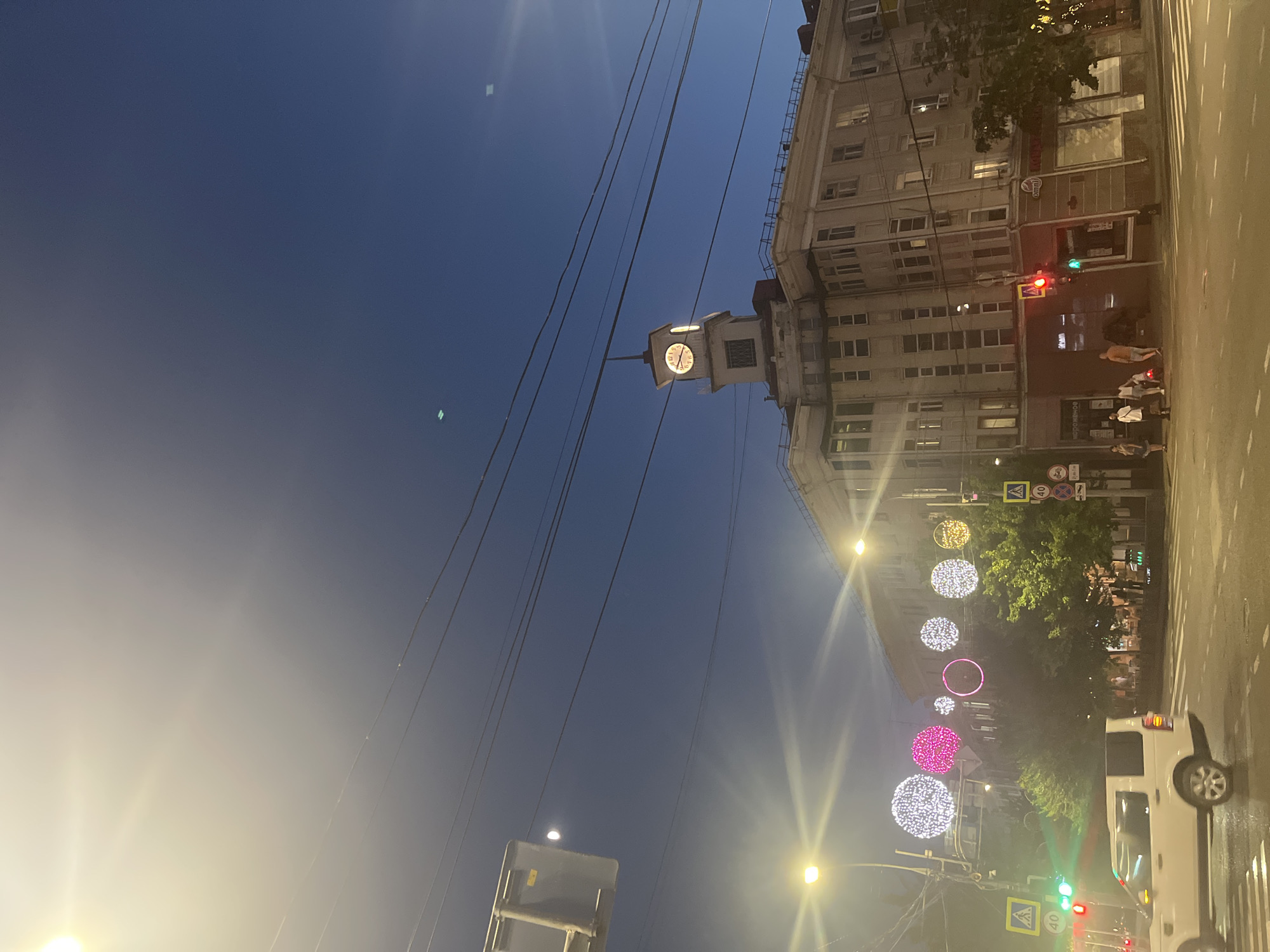 The image depicts a street scene at dusk in Krasnodar, Russia. The sky is a deep blue, transitioning to a lighter hue near the horizon, suggesting the sun has recently set. A large, ornate building dominates the right side of the frame, with multiple windows and a clock tower. The clock face is illuminated, showing the time.
The street is lined with trees and decorated with large, glowing spherical lights in various colors, including white, pink, and yellow. These lights are strung along the street, creating a festive atmosphere. A white SUV is visible on the road, along with several pedestrians walking on the sidewalk. Traffic lights are also present, showing red and green signals.
Several signs with text are visible, including road signs with numbers and symbols, and some lettering on the building facade. The overall mood is one of urban twilight, with a touch of festive celebration due to the decorative lights. The lighting suggests it's a cool evening.
