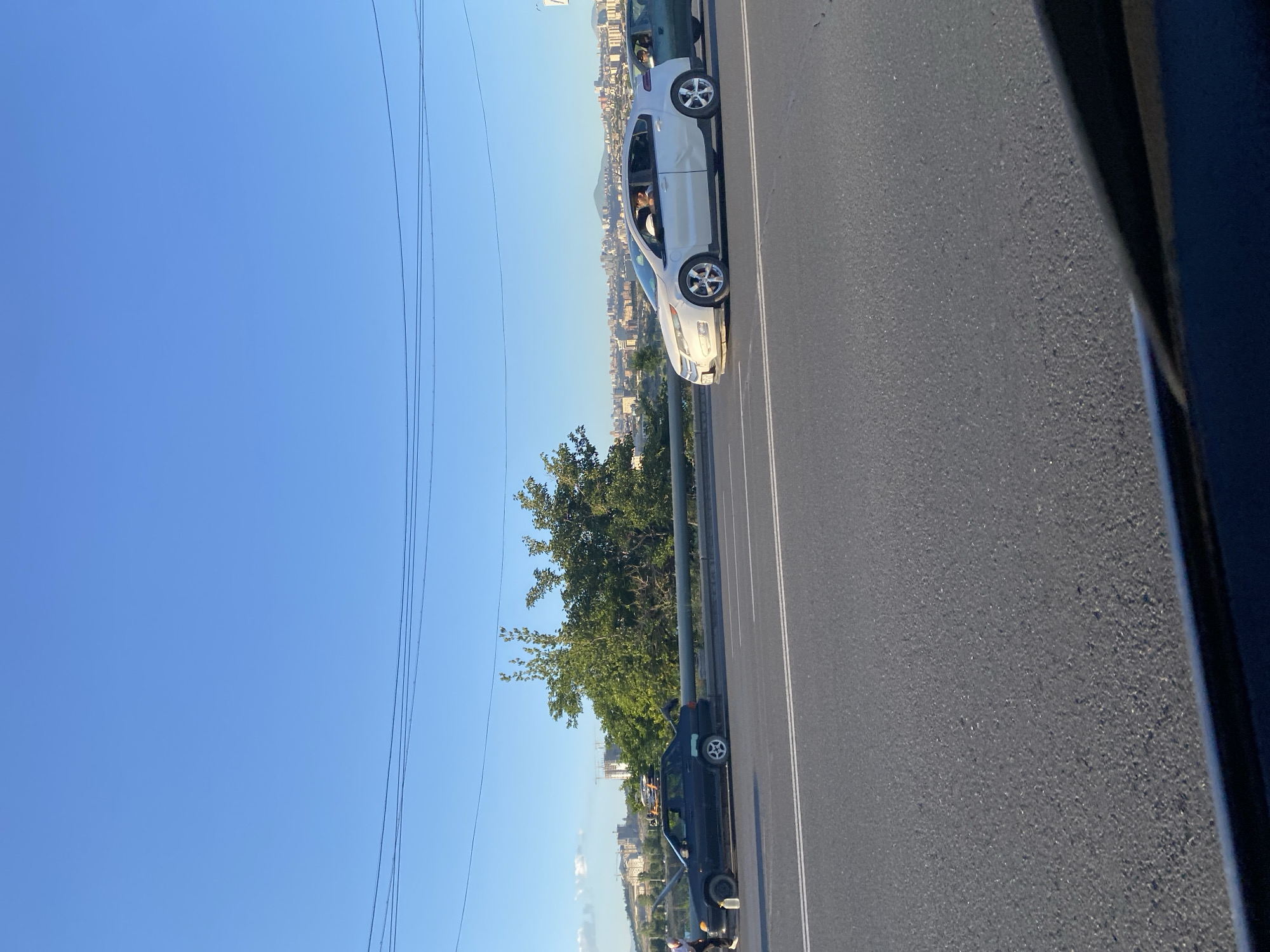 A black SUV is parked on the side of a road, with a person or people in the driver's seat. A white car is driving on the road, with a person in the driver's seat. The road is paved with asphalt and has white lane markings. There are trees and buildings in the background. The sky is clear and blue, suggesting it is daytime. The scene is set in Yerevan, Armenia.