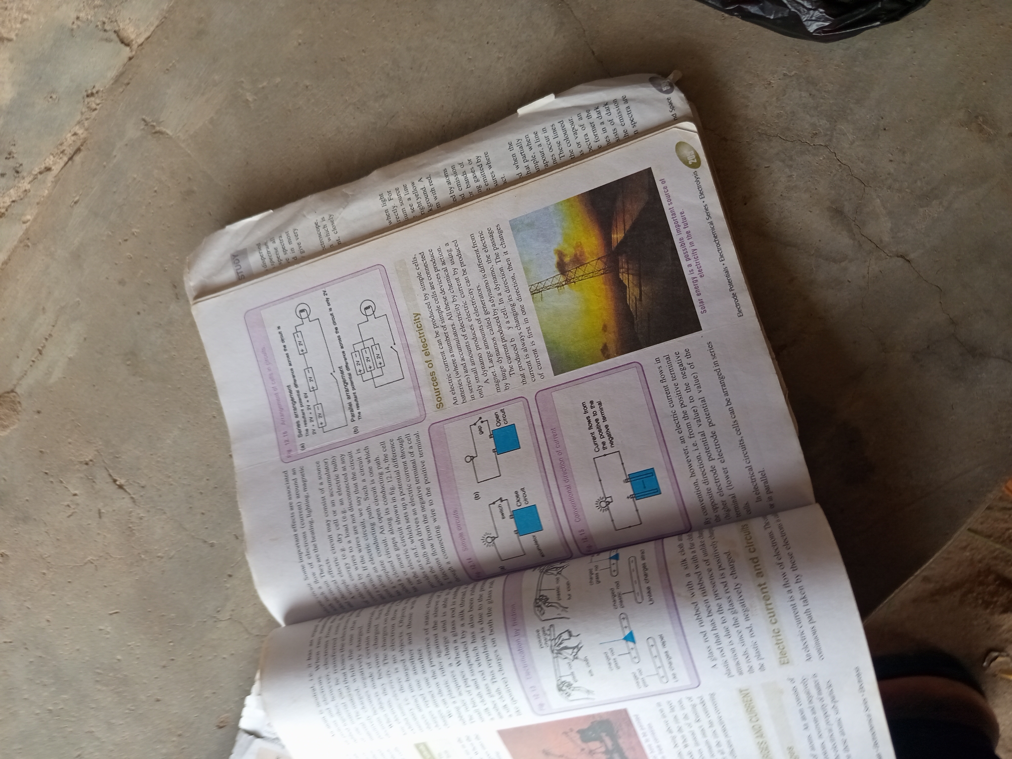 The image shows an open textbook with pages displaying text and diagrams related to electricity. One page features an image of a high-voltage electricity pylon against a colorful sky, alongside a diagram illustrating electrical circuits and components like batteries and switches. The textbook is laid open on a rough, concrete-like surface. The lighting suggests an indoor or shaded outdoor environment. The scene is static, with no people or discernible actions taking place within the frame.