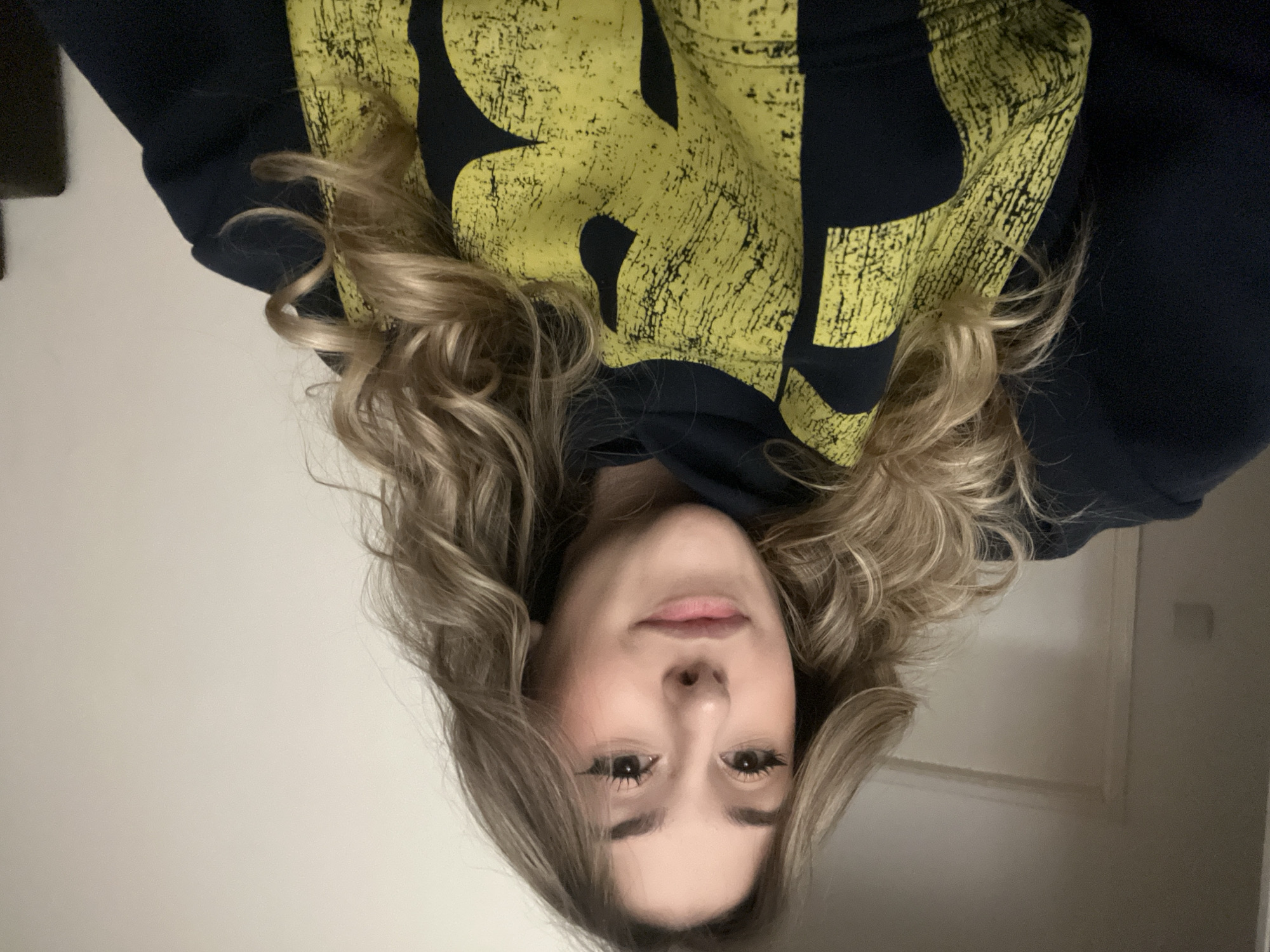 A fair-skinned young woman is centered in the image, with her face filling the bottom third of the frame. She has light blonde, wavy hair framing her face. She is wearing a dark blue hooded sweatshirt with a distressed yellow graphic on the hood. The background is a plain, light-colored wall. The image is upside down. There is no indication of Sofia, Bulgaria.