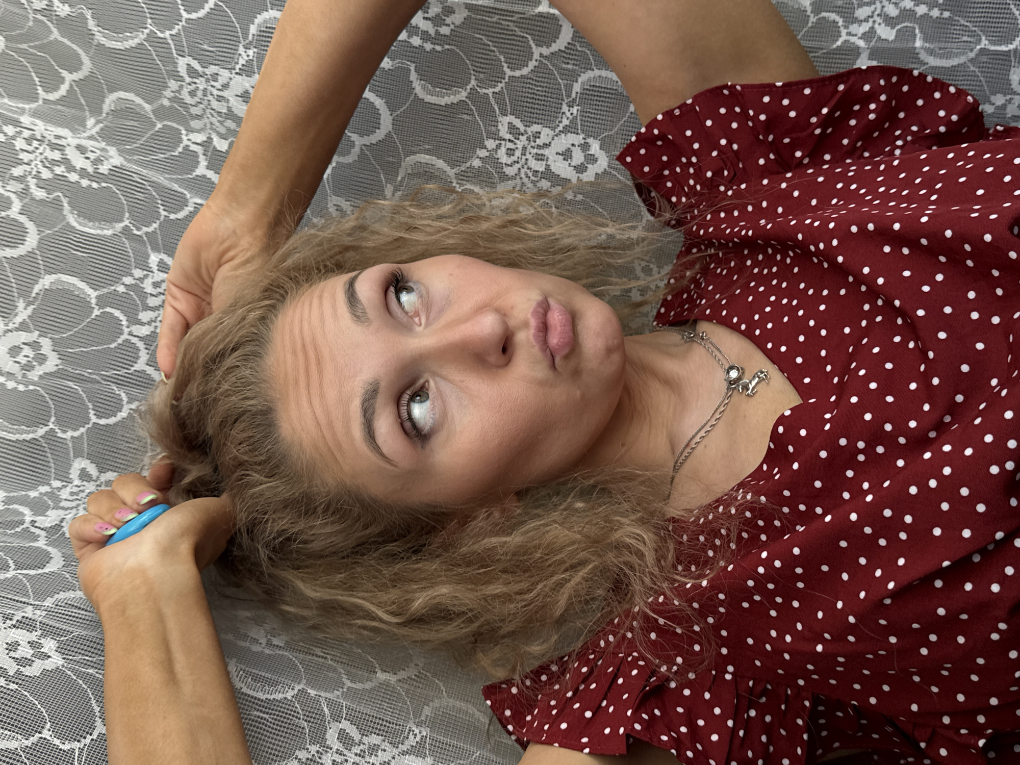 A woman is lying on a patterned white and grey surface, possibly a bed or tablecloth. She is wearing a red polka-dot dress and a silver necklace. Her curly blonde hair is spread around her head. Her right arm is bent with her hand holding a blue and green object, while her left arm is raised above her head. She is looking upwards with a pouting expression. The location and city of Chaikovsky, Russia, are not deducible from the visual information provided.