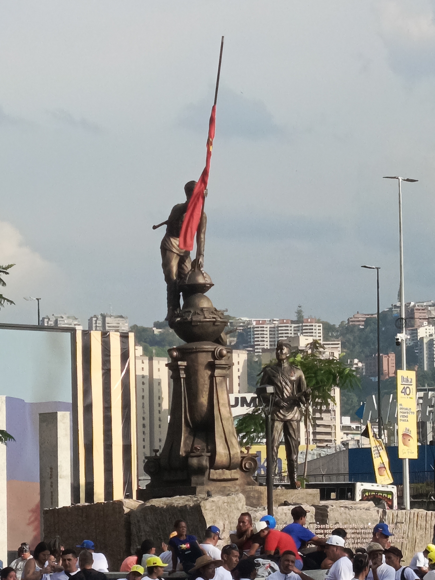 The image depicts a public square or plaza in Caracas, Venezuela, during daytime under an overcast sky. Central to the scene is a large, bronze-colored monument. The primary statue on top features a strong male figure holding a prominent red flag that extends upwards. Below this, another smaller statue of a man in uniform stands at the base of the monument.
Around the monument's base, a diverse crowd of people, mostly men, are gathered. Some are sitting on the stone structure, while others stand nearby, suggesting a public assembly, rally, or social gathering. Many individuals are wearing baseball caps or hats.
In the background, the city of Caracas stretches across the landscape, with numerous multi-story buildings and residential structures climbing up green, rolling hills. Streetlights punctuate the scene. On the left, part of a large billboard or urban art installation with abstract shapes and black and yellow stripes is visible. On the right, a bright yellow sign clearly displays "Daka 40" and fragmented Spanish text which appears to be "EL REGALO PERFECTO" (The Perfect Gift). Other partially legible text fragments, such as "UM," "Unlo," "bvialitosnia," "Marable sacr," and "Guerra Palt" are also visible on surrounding structures. The overall impression is one of a vibrant urban space hosting a community event.