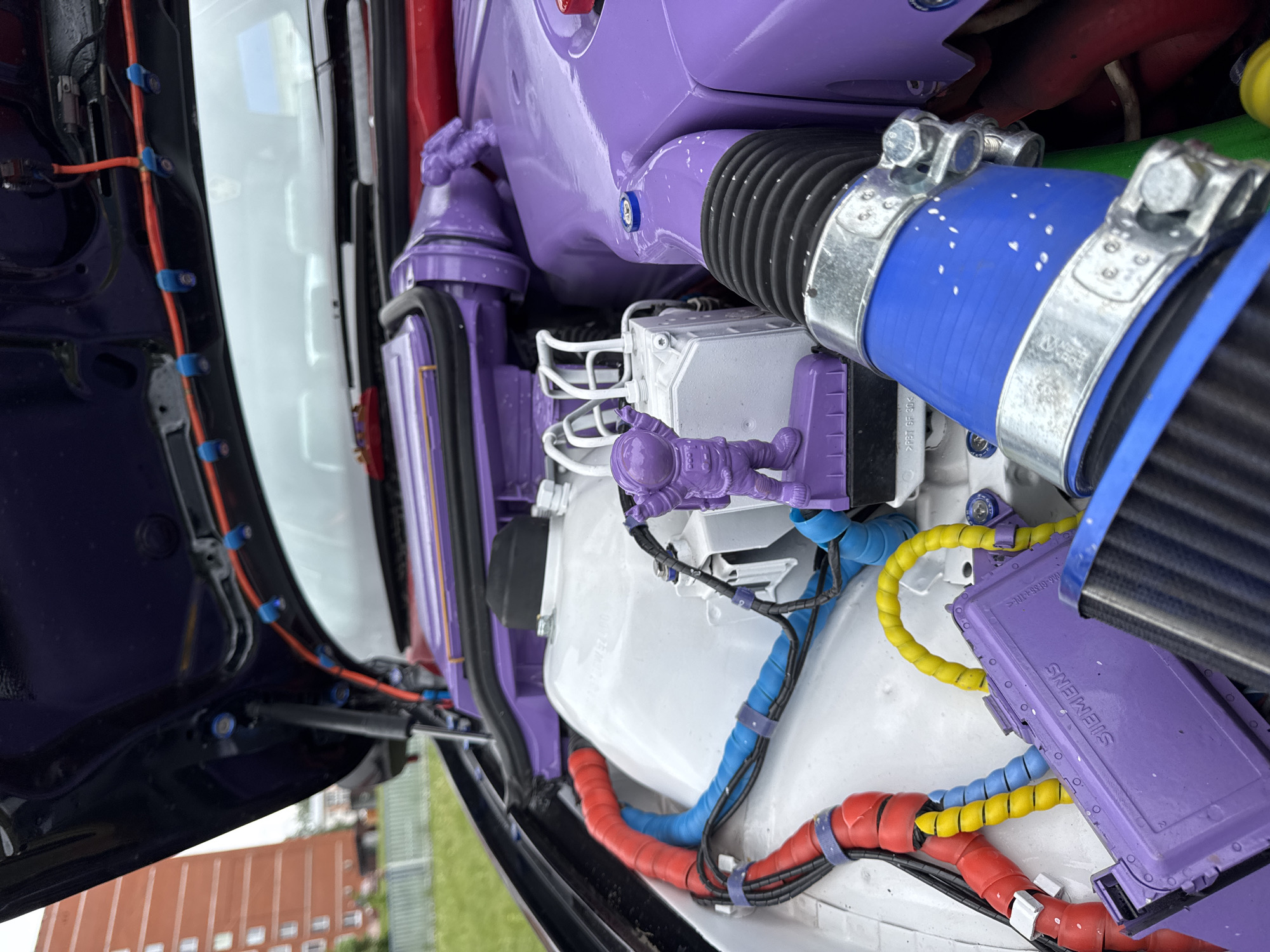 A close-up shot of a customized engine compartment, rotated to give a vertical perspective, reveals an array of components in a vibrant purple, white, blue, red, and yellow color scheme. The visible wires and tubes are neatly organized and color-coded. A small, purple astronaut figurine is affixed to a white component. The branding "SIEMENS" is visible on a purple module. Hoses are secured with silver clamps. The hood is open, revealing a dark blue inner panel with orange wiring. The background shows an out-of-focus landscape, including the top of a building with multiple windows and greenery, which suggests the city of Chaikovsky, Russia.