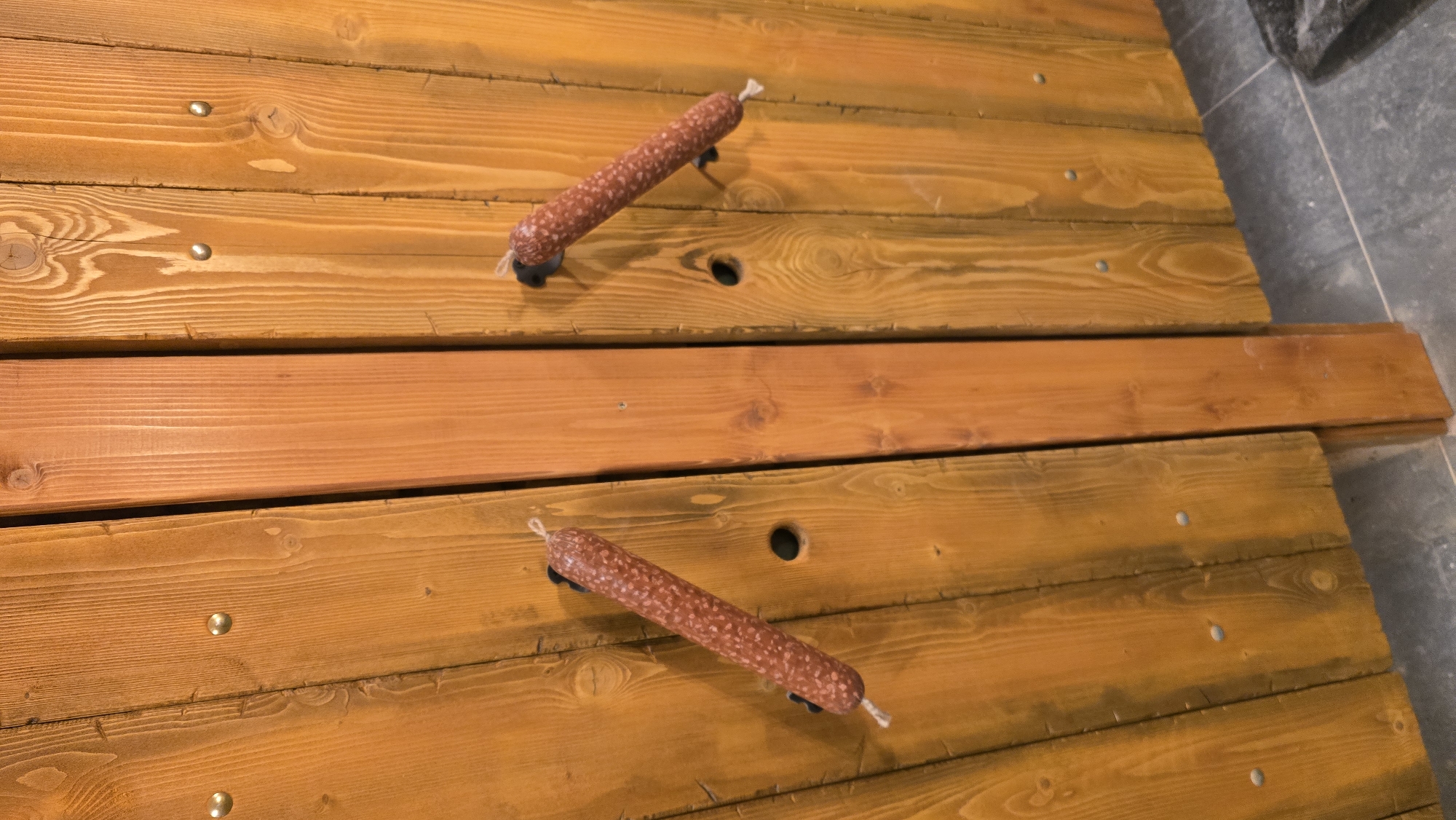 A high-angle shot shows two salamis, each with black cylindrical supports, on a wooden surface comprised of planks in Porech'e, Russia. The wood is varnished with a light brown stain. Silver studs are visible on some planks. One larger plank runs horizontally across the center. A portion of a gray, tiled floor is visible in the upper right and lower right corners.