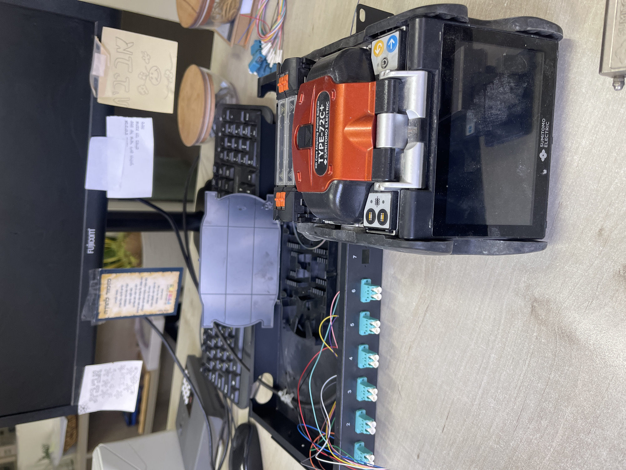 The image depicts a work desk with a computer monitor in the background. Attached to the monitor are several notes in Hebrew. On the desk, a fiber optic splicing machine, identified as a "SUMITOMO ELECTRIC TYPE-72C+", is prominently displayed. Next to it, a fiber optic distribution box with several colorful fiber optic cables and connectors is visible. A keyboard and mouse are also present on the desk. The overall environment suggests an office or technical workspace. The image does not contain any specific visual cues that would allow for precise localization within Tel Aviv, Israel.