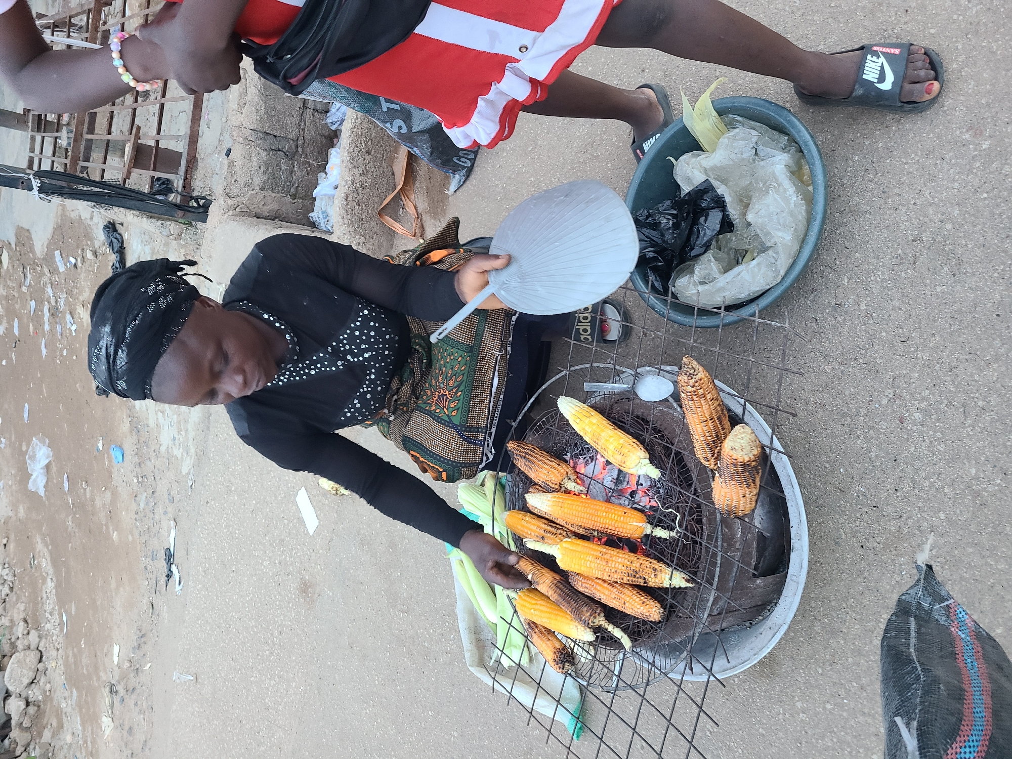 In Jos, Nigeria, a woman is seen grilling corn on a charcoal stove outdoors. She wears a black headscarf and a patterned skirt. A younger person, possibly a girl, stands next to her, wearing a red dress. Several ears of corn are arranged on a wire grill over the glowing coals, with some already charred. In the foreground, a grey plastic basin contains plastic bags and some corn husks. The ground is paved with concrete. To the left, a black mesh bag is visible. The scene appears to be a street-side food stall.