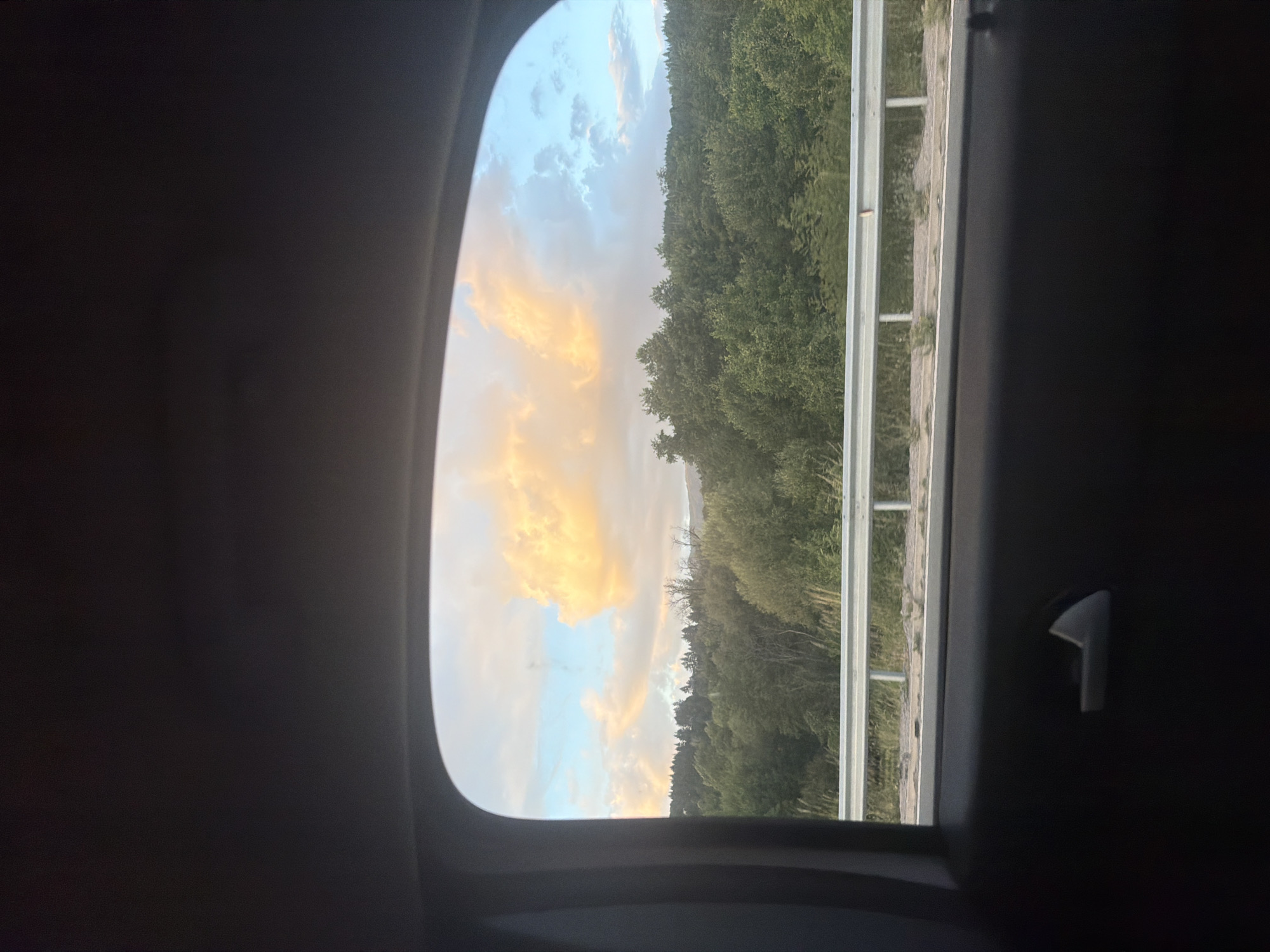 The image captures a view from inside a vehicle looking out of a rear window at a rural landscape. The window frame is dark and partially obstructs the view. In the distance, a dense forest covers rolling hills, with trees of varying shades of green. A section of a guardrail, appearing to be made of metal, runs horizontally across the middle ground. Beyond the guardrail, there is a glimpse of what seems to be a road or embankment with some vegetation. The sky above is partially filled with clouds; on the left, large, puffy clouds are illuminated with a warm, golden-orange light, suggesting either sunrise or sunset. On the right, the sky transitions to a pale blue with scattered white clouds. The image was taken in Studena, Bulgaria.