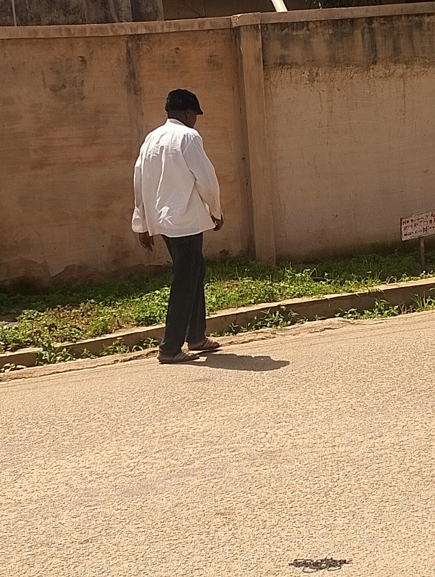 The image depicts a man walking away from the camera on a paved road. He is wearing a white long-sleeved shirt, dark trousers, a dark cap, and sandals. He is walking along a sidewalk bordered by a concrete curb and a patch of green vegetation. Behind the vegetation is a textured, light-colored wall, possibly a building or boundary wall, with some staining and discoloration. A small sign with writing is attached to the wall on the right side of the image. The overall scene appears to be outdoors in a location that could be a street or pathway in Jos, Nigeria, given the environmental context of the wall, vegetation, and road surface. The lighting suggests it is daytime, with shadows cast on the road.