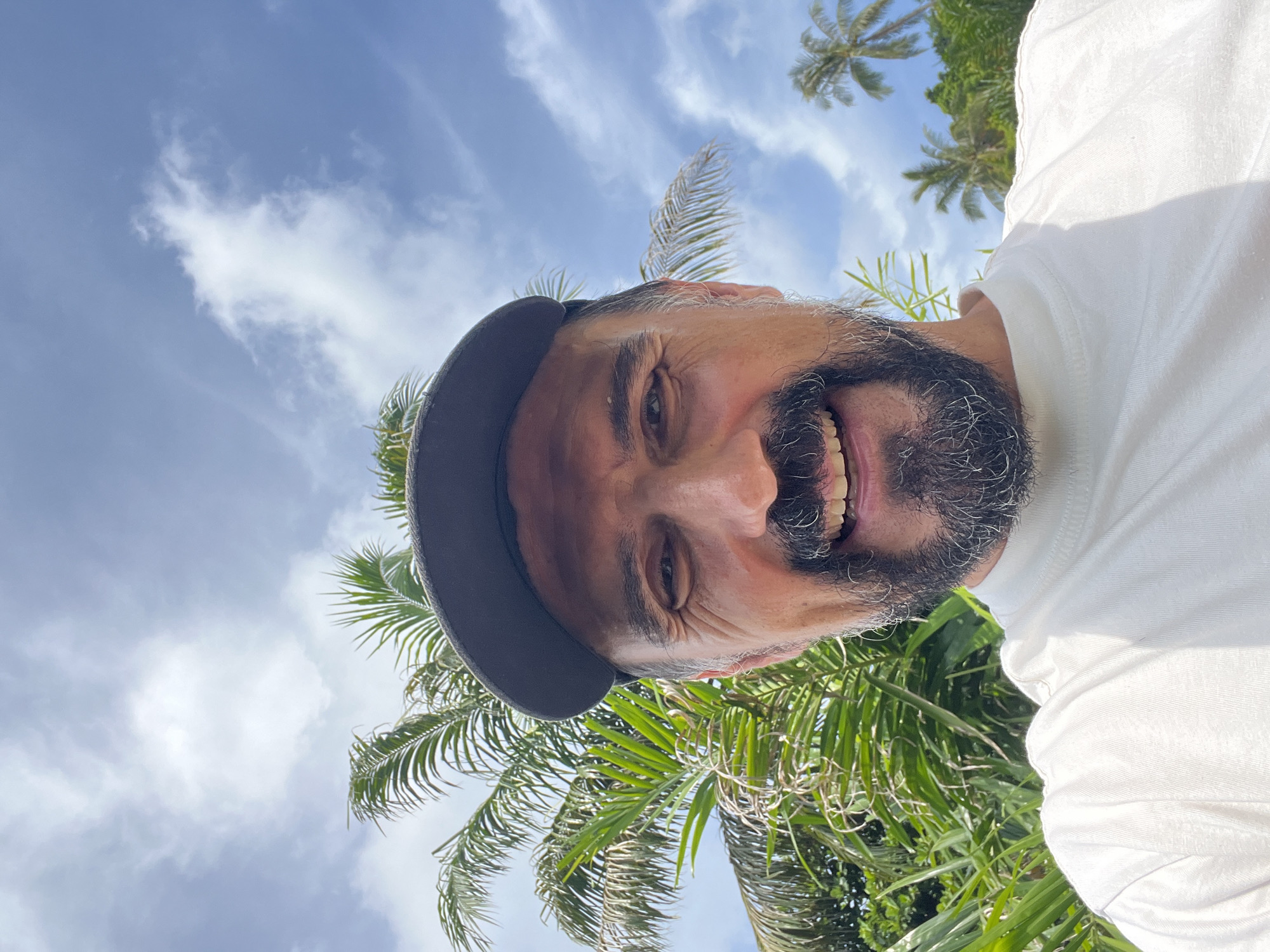 A close-up selfie of a middle-aged man wearing a white t-shirt and a dark baseball cap is taken outdoors. He has a beard and mustache, and he is smiling widely, showing his teeth. The background consists of lush green palm trees and a bright blue sky with scattered white clouds, indicative of a tropical location like Baan Chaweng Noi, Thailand. The lighting suggests it is daytime with sunlight casting shadows.