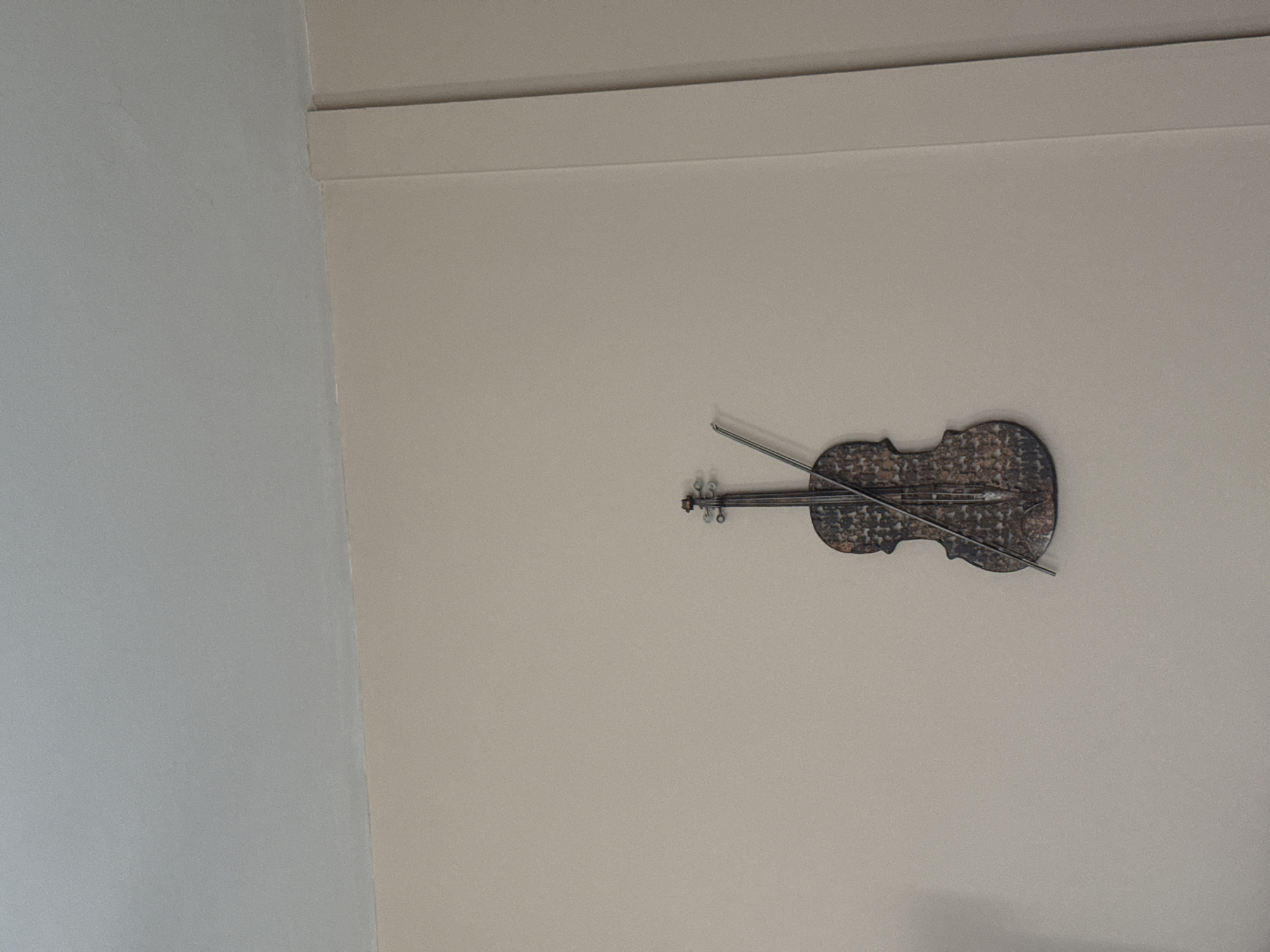 The image shows a decorative metal violin and bow hanging on a beige wall. The violin is horizontally oriented, with the bow resting across the body of the instrument. A white wall is visible in the left part of the image. The image does not provide information to determine a location in Sofia, Bulgaria.