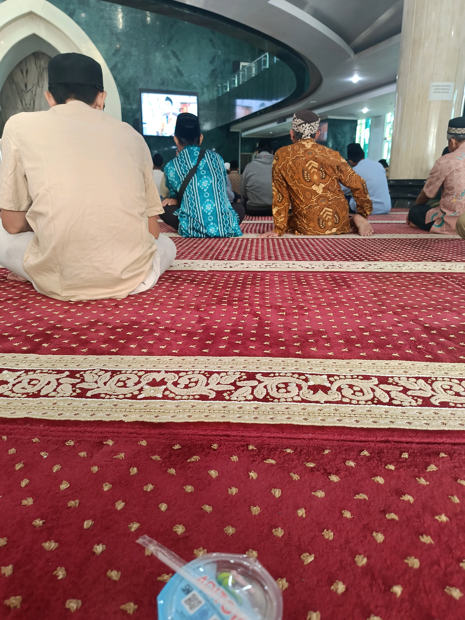 The image depicts an interior view of a mosque in Banguntapan, Indonesia, with several individuals seated on a red carpet during what appears to be a religious gathering. The carpet features a repeating pattern of small gold motifs and a decorative border. In the foreground, a clear plastic cup with a straw is visible. In the midground, individuals are seated in rows, facing away from the camera. One person in a tan shirt and black cap is on the left. To their right, a person in a turquoise patterned garment and black cap is also seated. Further to the right, a person in a brown patterned garment and a patterned head covering is visible. Behind them, other individuals are seated, with some wearing light blue and patterned shirts. A television screen displaying an image is mounted on the wall in the background, and the mosque's architecture, including a curved ceiling and pillars, is discernible. The overall scene suggests a prayer or lecture session taking place within the mosque.