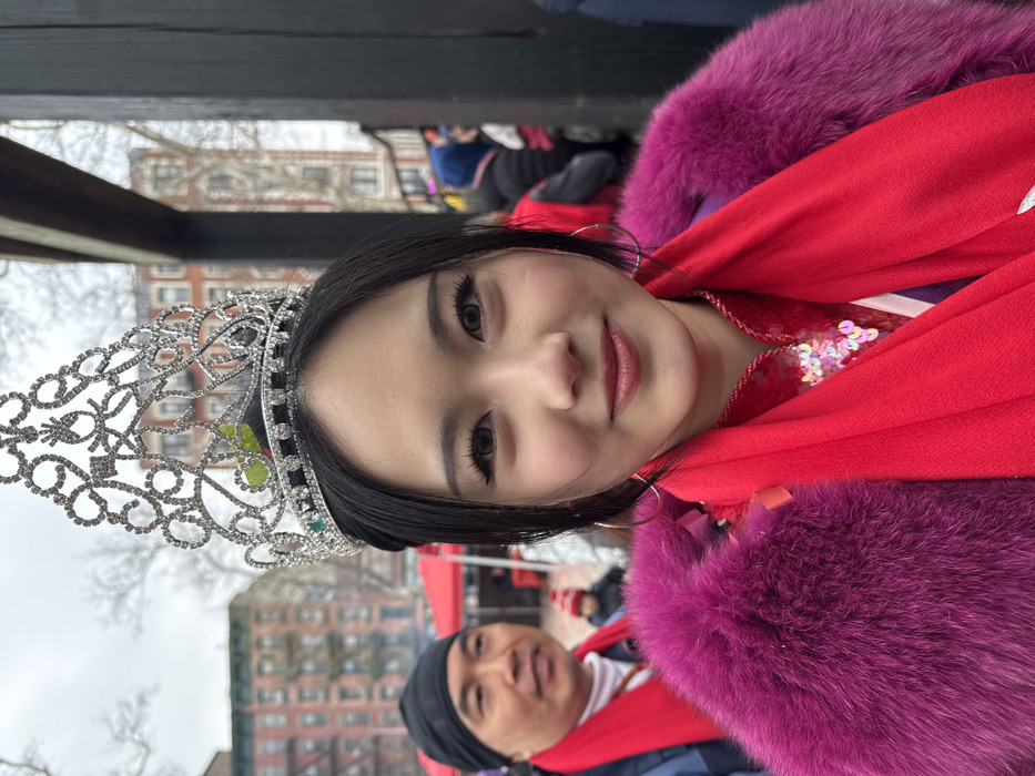 A woman with dark hair wears a silver crown, hoop earrings, and a bright red scarf with magenta fur trim. She smiles slightly at the camera. A man in a black beanie is visible in the lower center of the frame, also wearing red. Buildings and trees are blurred in the background, suggesting an outdoor location. The sky appears overcast. The overall impression is of a festive or celebratory event.