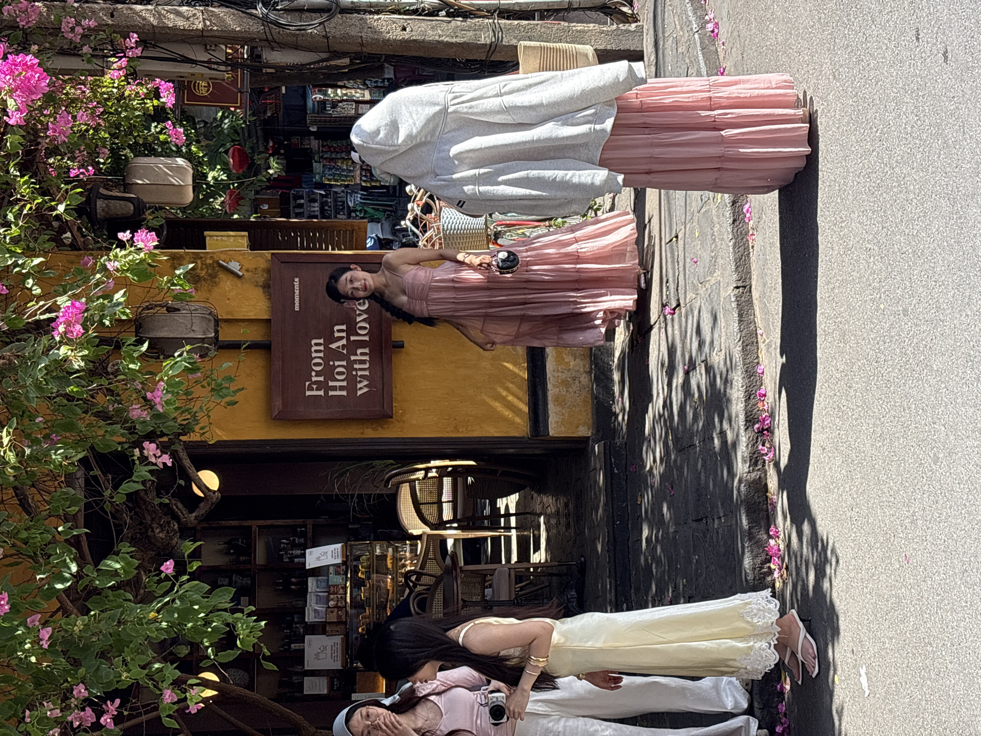 In Hội An Ward, Vietnam, on a sunny day, three individuals are present on a street with a building facade. The building has a yellow-painted exterior and a wooden sign that reads "From Hoi An with love" and "moments". Above the sign and extending from the building are branches of a tree with green leaves and pink flowers, with decorative lanterns hanging from them.
On the left, two young women are standing. One, with a light blue headscarf, wears a light pink top and white pants and holds a camera. The second woman wears a light yellow, floor-length dress with intricate white lace detailing around the hem and white heeled flip-flops.
In the center, a woman in a pink, tiered, floor-length dress stands facing forward. She is holding a small, round, black bag with a chain strap in her right hand.
On the right, a person is standing with their back to the camera, wearing a grey hooded sweatshirt over a pink, tiered, floor-length skirt. This individual also has a straw bag hanging from their shoulder.
The street in front of the building is paved with dark stone tiles, and fallen pink flower petals are scattered on the ground. The ground also shows dappled sunlight and shadow patterns from the overhead foliage. In the background, the interior of the building appears to be a shop with shelves stocked with bottles and other items, and wicker chairs are visible.