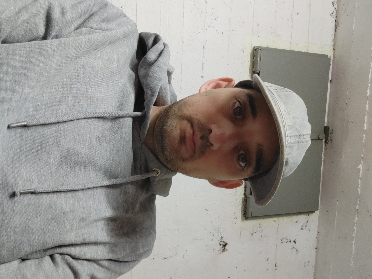 A man with dark hair and a beard is wearing a gray hoodie and a gray baseball cap. He is looking at the camera. Behind him is a white concrete wall with peeling paint. A gray metal door is on the wall. This does not have enough information to be connected to the city of Tel-Aviv, Israel.